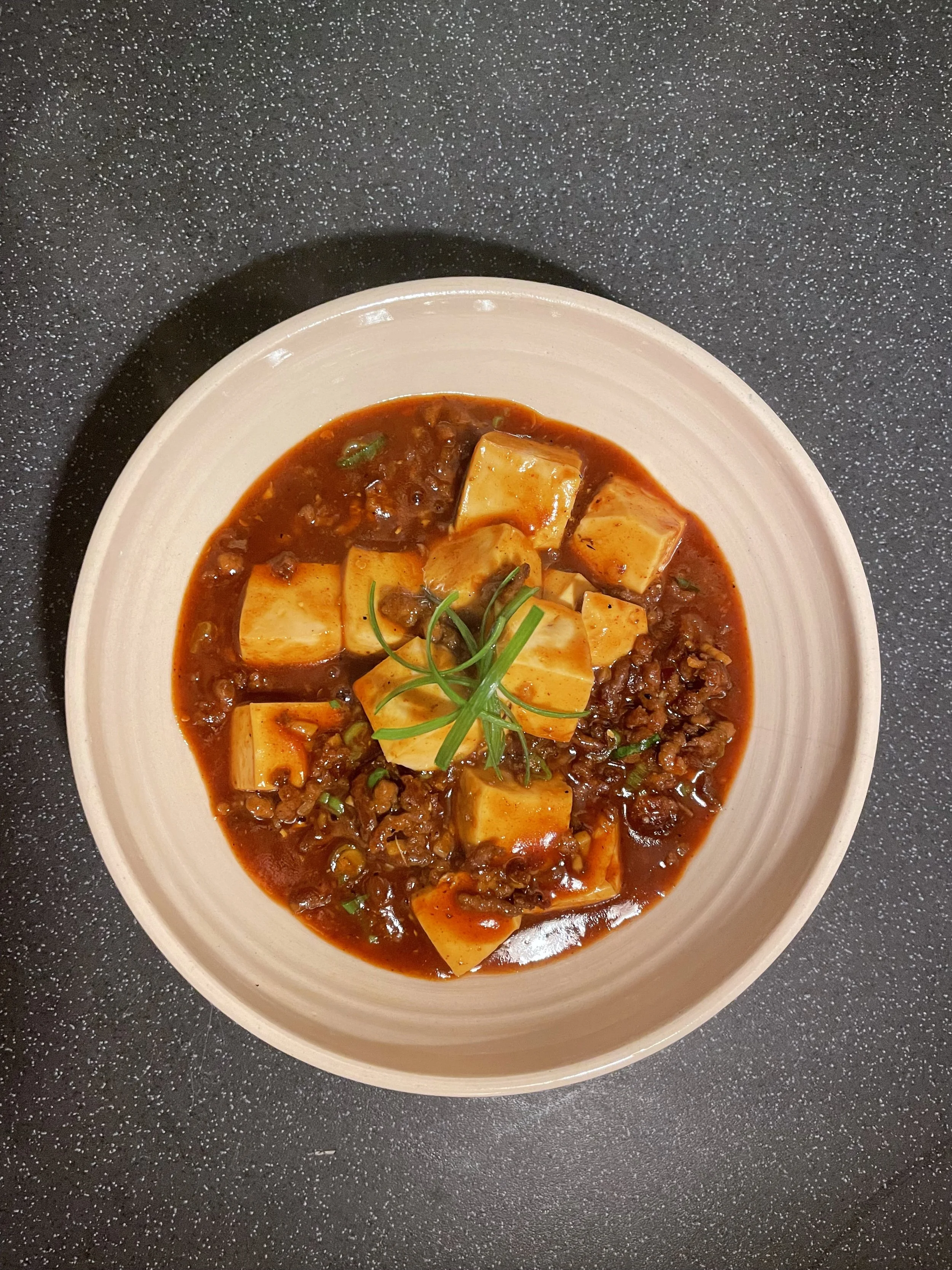 Bowl of spicy tofu and ground meat stew garnished with green herbs on a dark speckled surface.