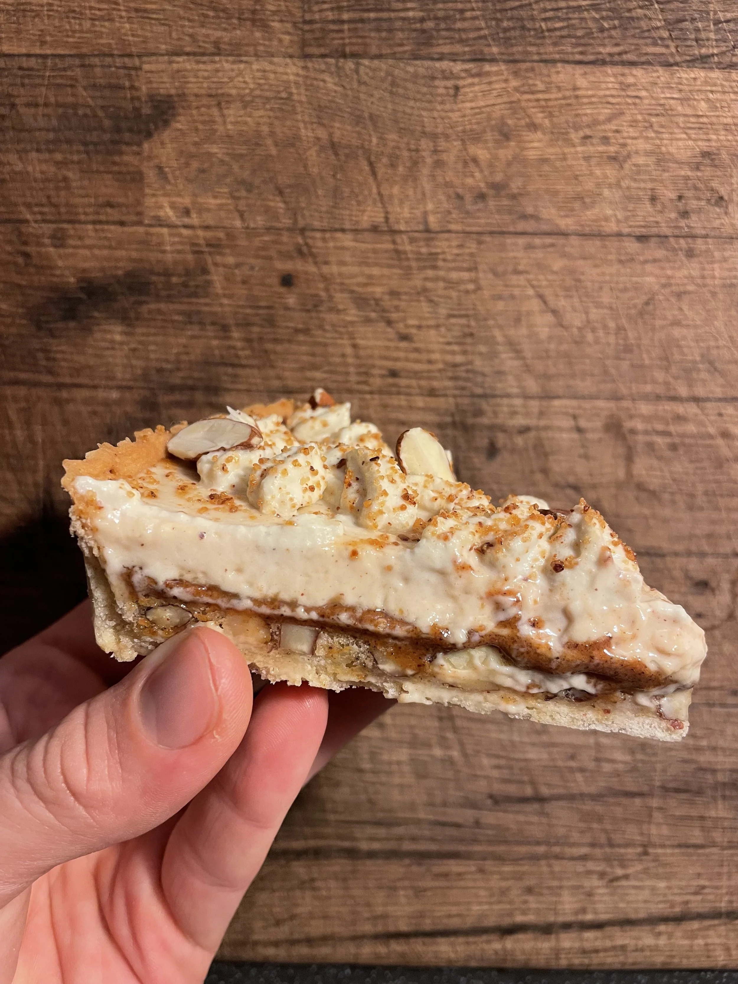 A hand holding a slice of pie with a creamy filling topped with nuts on a wooden surface.