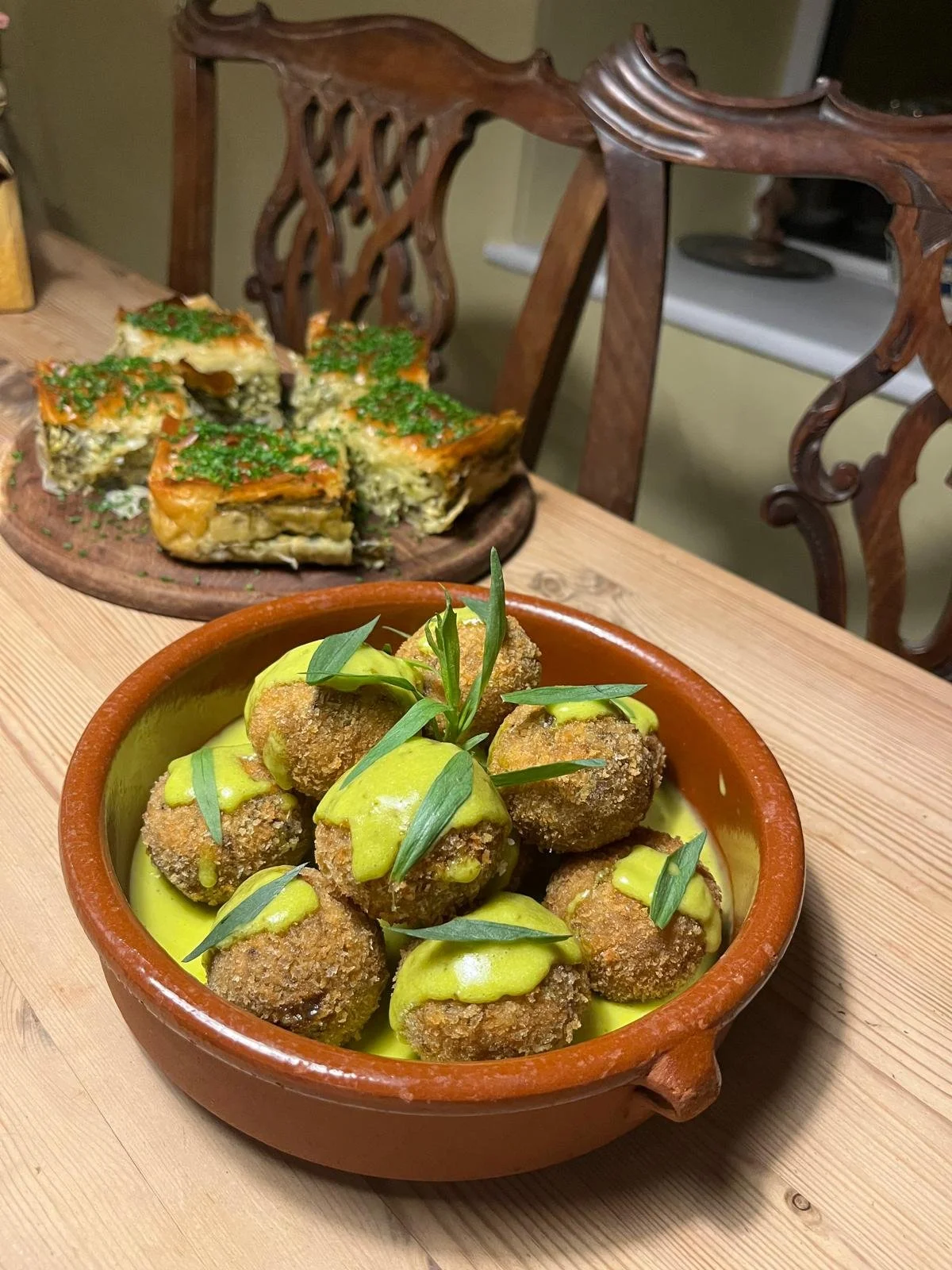 A bowl of assorted breaded fried appetizers garnished with green sauce and herbs, with a plate of vegetable quiche or casserole topped with chopped herbs in the background on a wooden table.