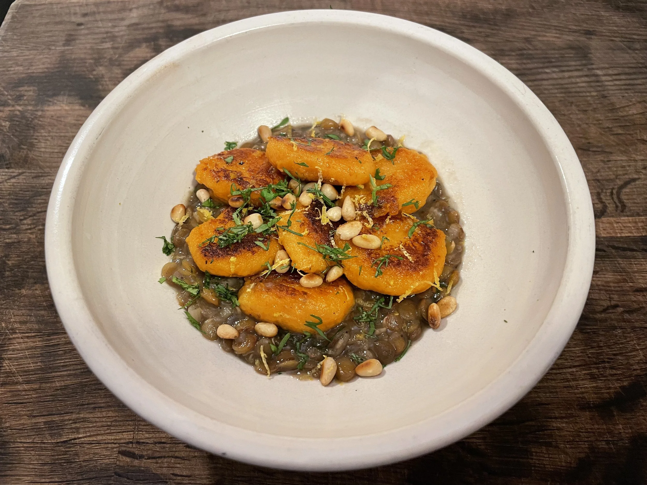 A bowl of roasted plantains garnished with chopped herbs and pine nuts, served over a bed of lentils on a wooden surface.