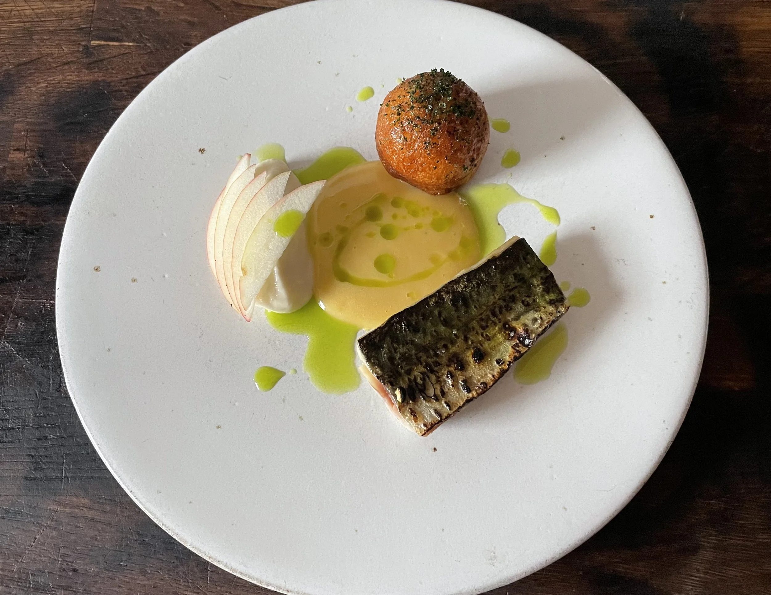 A white plate with a grilled fish fillet, a turnip puree with green oil, a fried apple doughnut with herbs, and thin slices of apple. The plate is on a dark wooden surface.