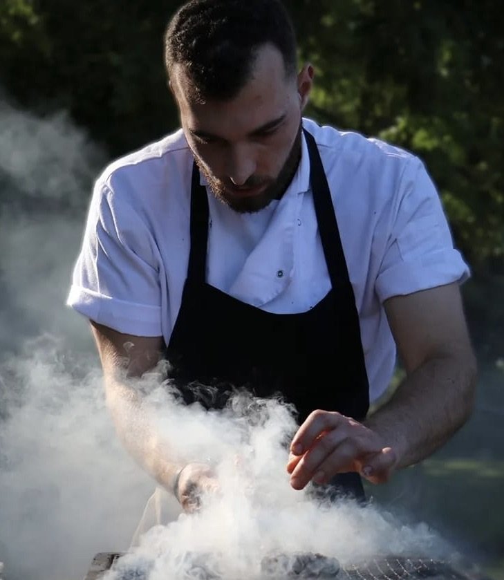 Chef cooking outdoors wearing a white shirt and black apron, grilling steaks on BBQ.