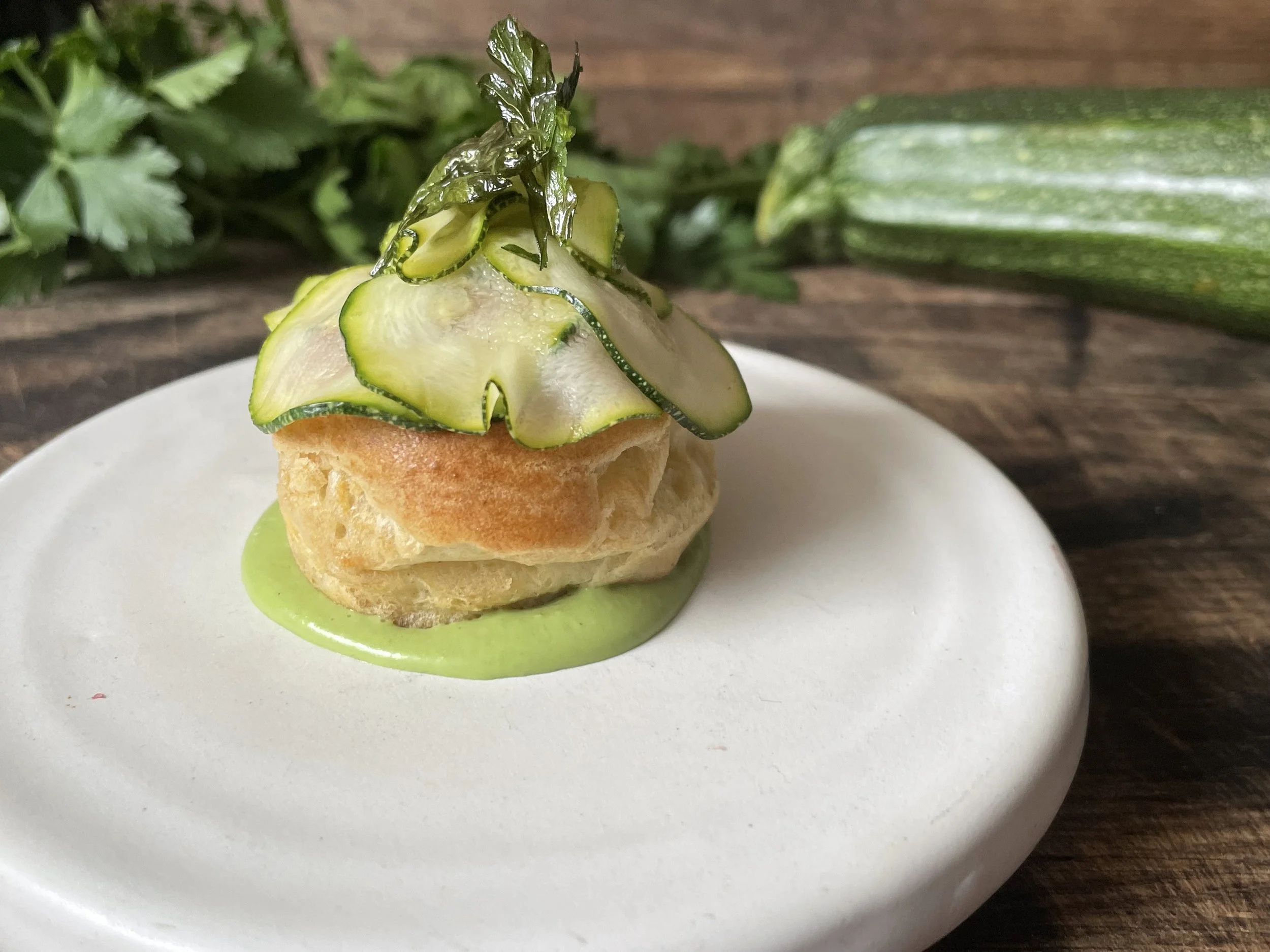 A small pastry topped with sliced cucumbers and a sprig of parsley, served on a white plate with green sauce underneath.