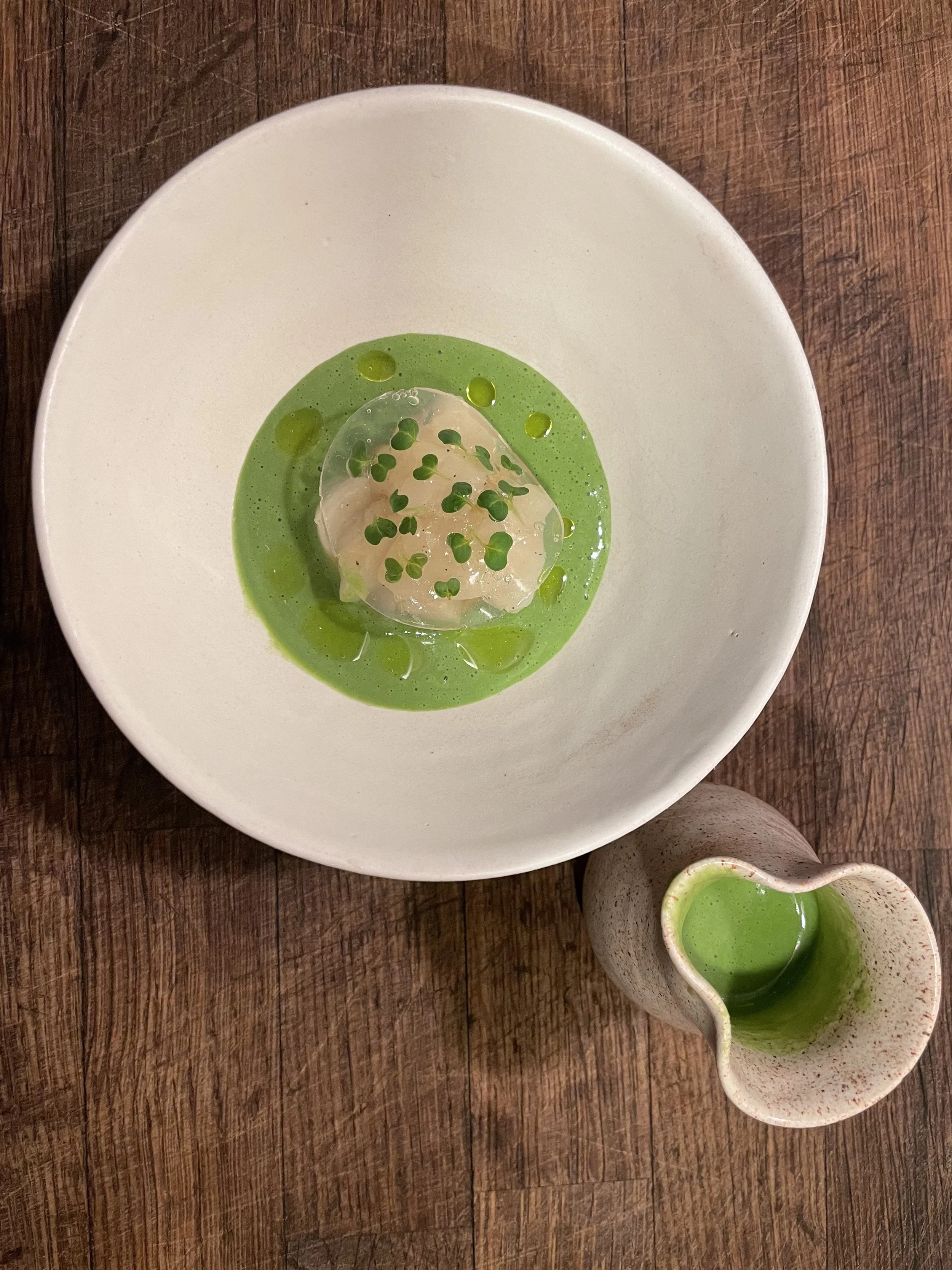 A white bowl with green sauce and a transparent gelatinous cube topped with microgreens, accompanied by a small pitcher of green liquid on a wooden surface.