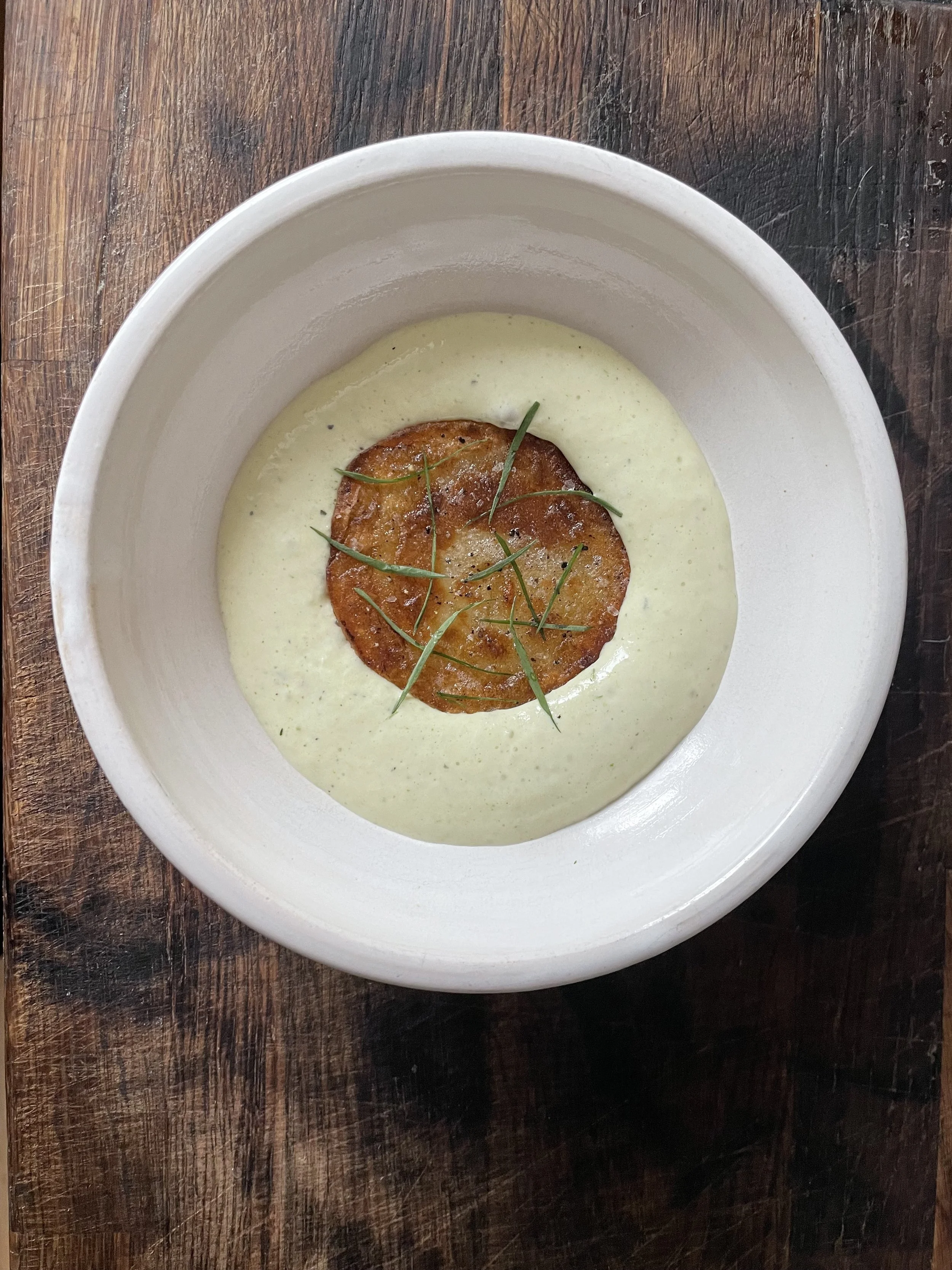 Creamy green soup garnished with a fried potato cake and thin chives in a white bowl on a wooden surface.