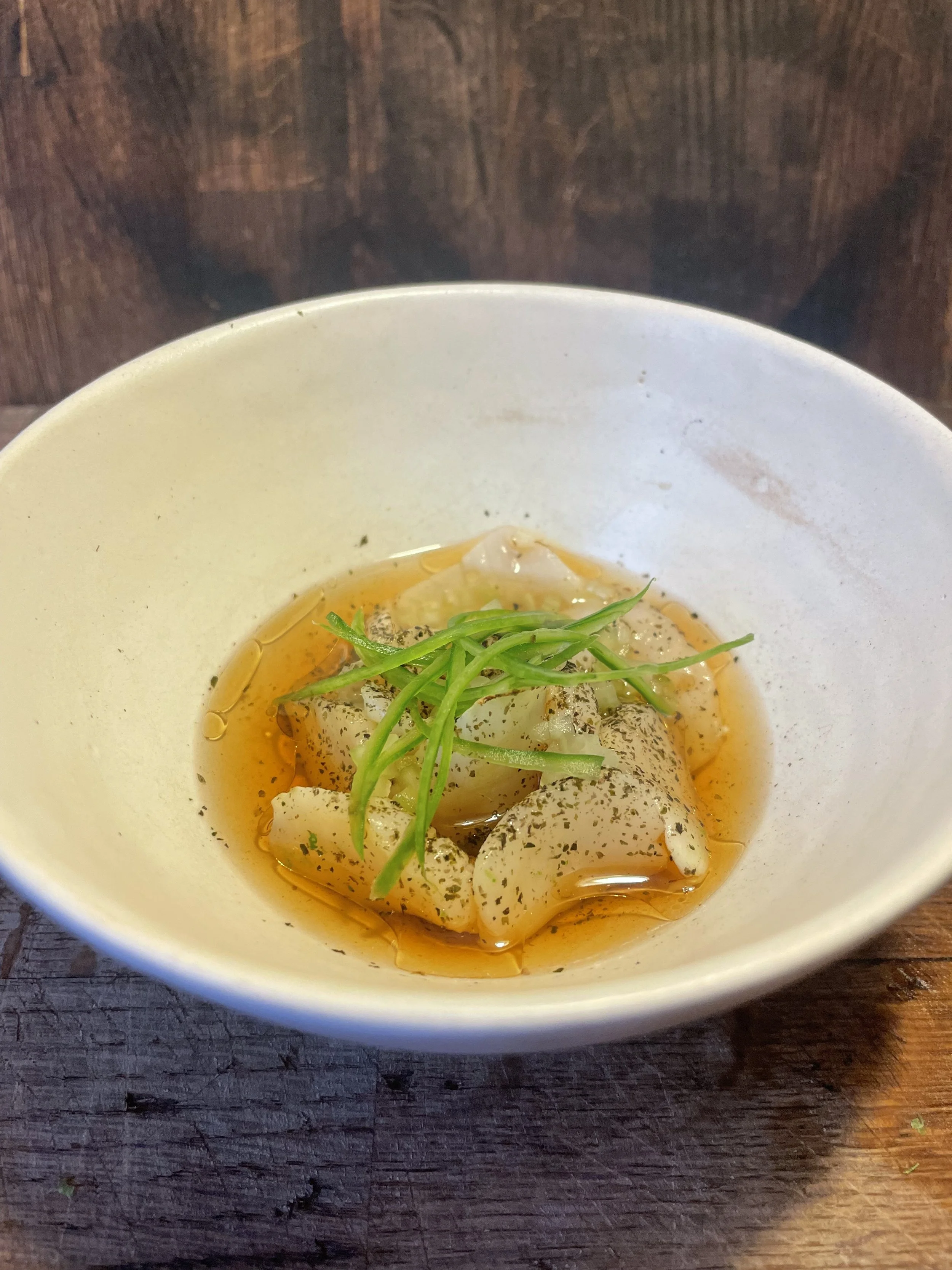 A bowl of thinly sliced fish or seafood garnished with green herbs, served in a light broth or sauce.
