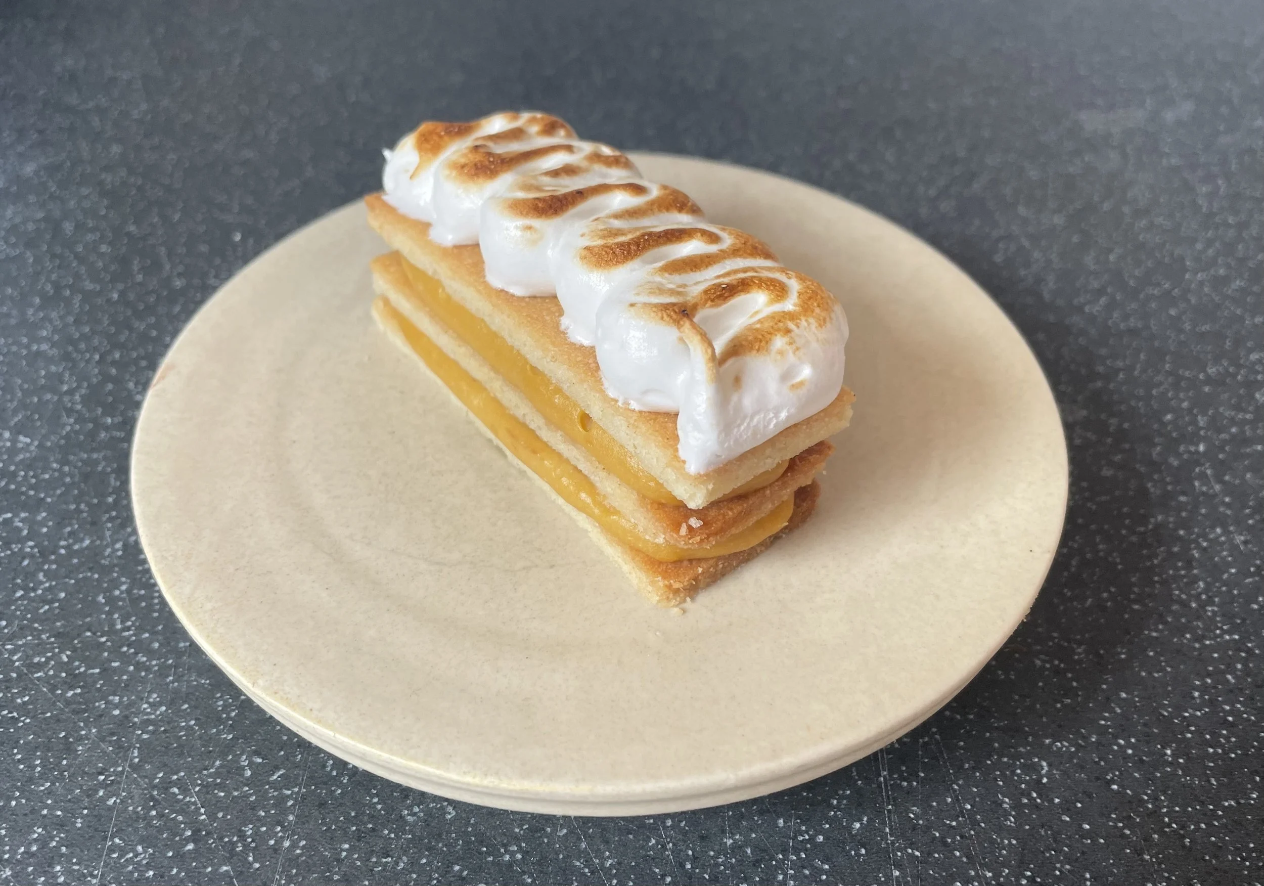 A mango Mille-Feuille dessert with meringue topping on a beige plate, on a dark speckled surface.