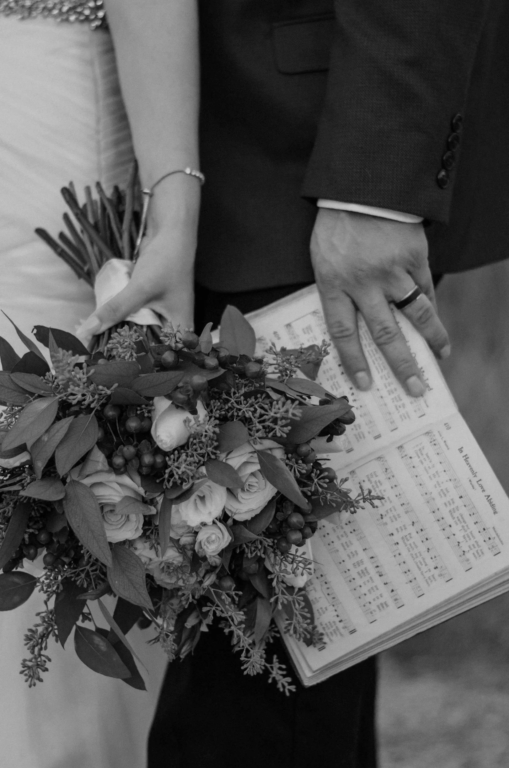 Close-up of a person holding a bouquet of flowers and sheets of music, wearing a suit with a wedding ring.