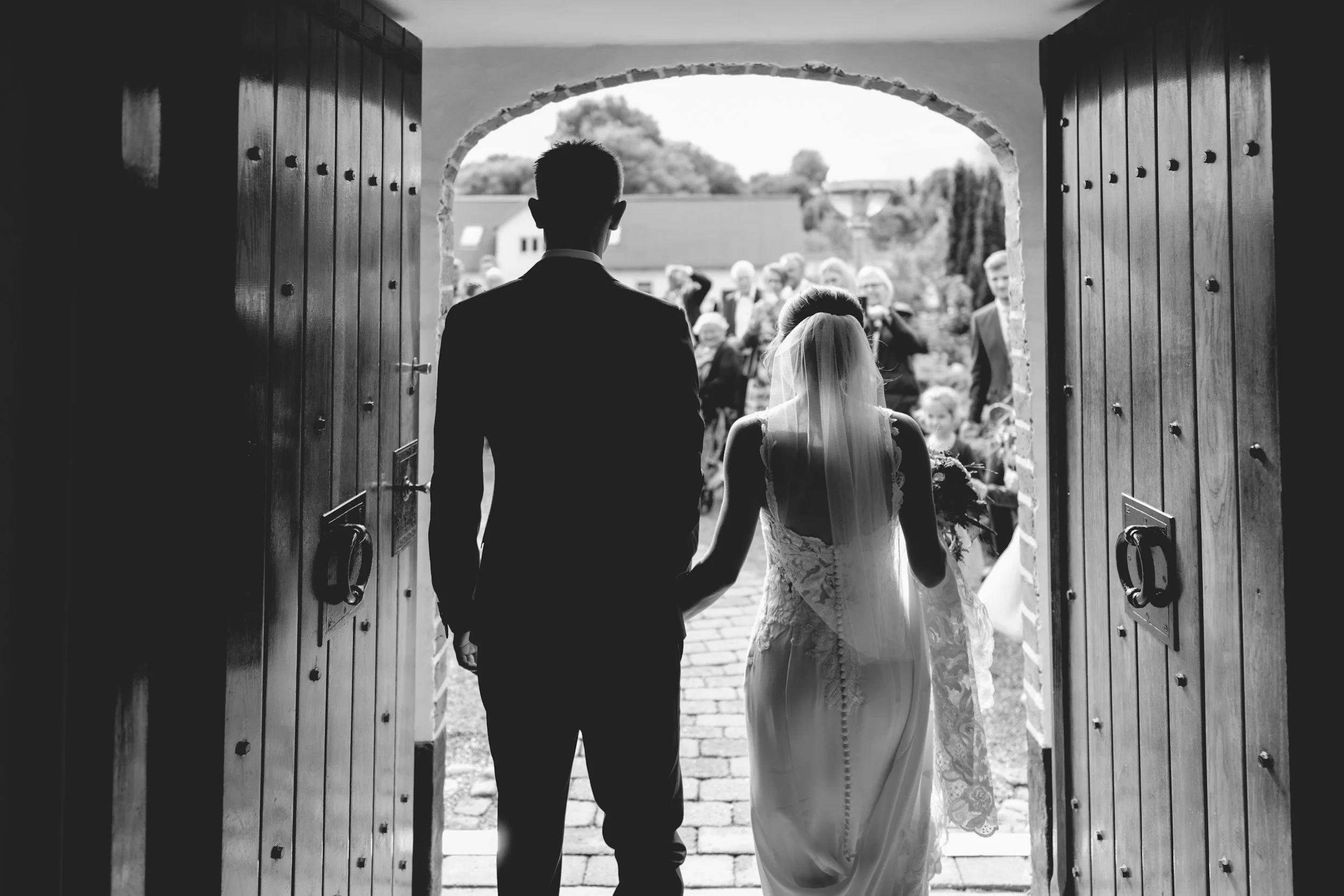 A bride and groom holding hands, walking out of a doorway towards a crowd of wedding guests outside.