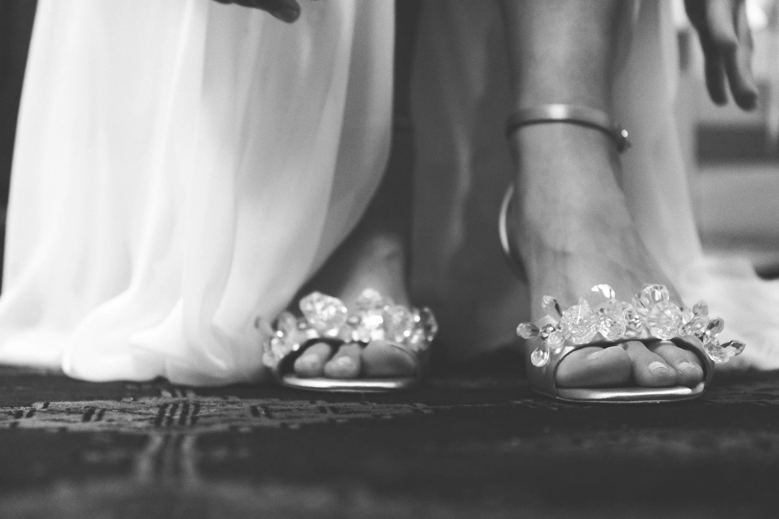 Close-up of a bride's feet wearing elegant sandals decorated with large gemstones, standing on a patterned rug, with her white dress flowing around her.