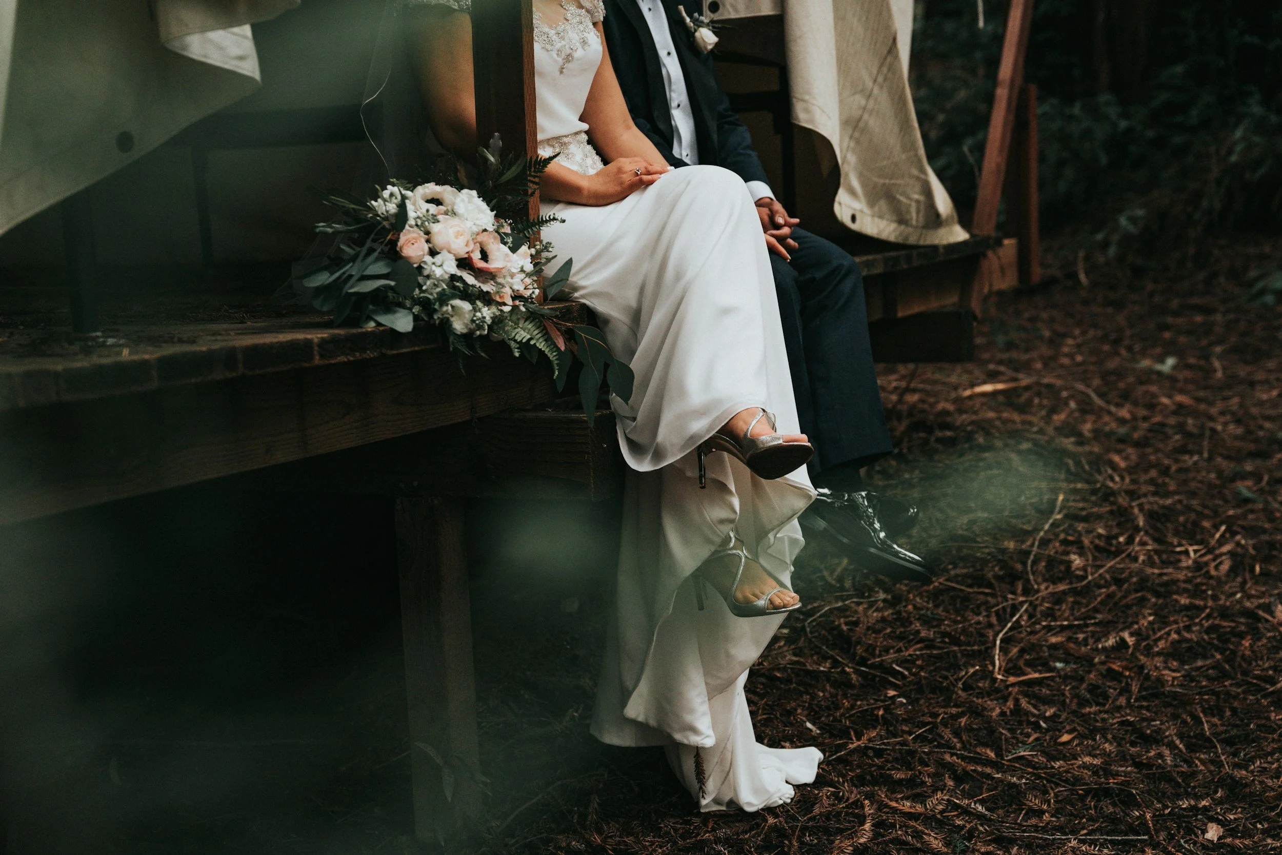 A bride and groom sit on a wooden platform outdoors; the bride wears a white dress, and the groom is in a dark suit. A bouquet of white and pink flowers lies next to them. Their legs and shoes are visible, with the bride wearing high heels and the groom in black shoes. The ground below is covered in brown leaves or mulch.