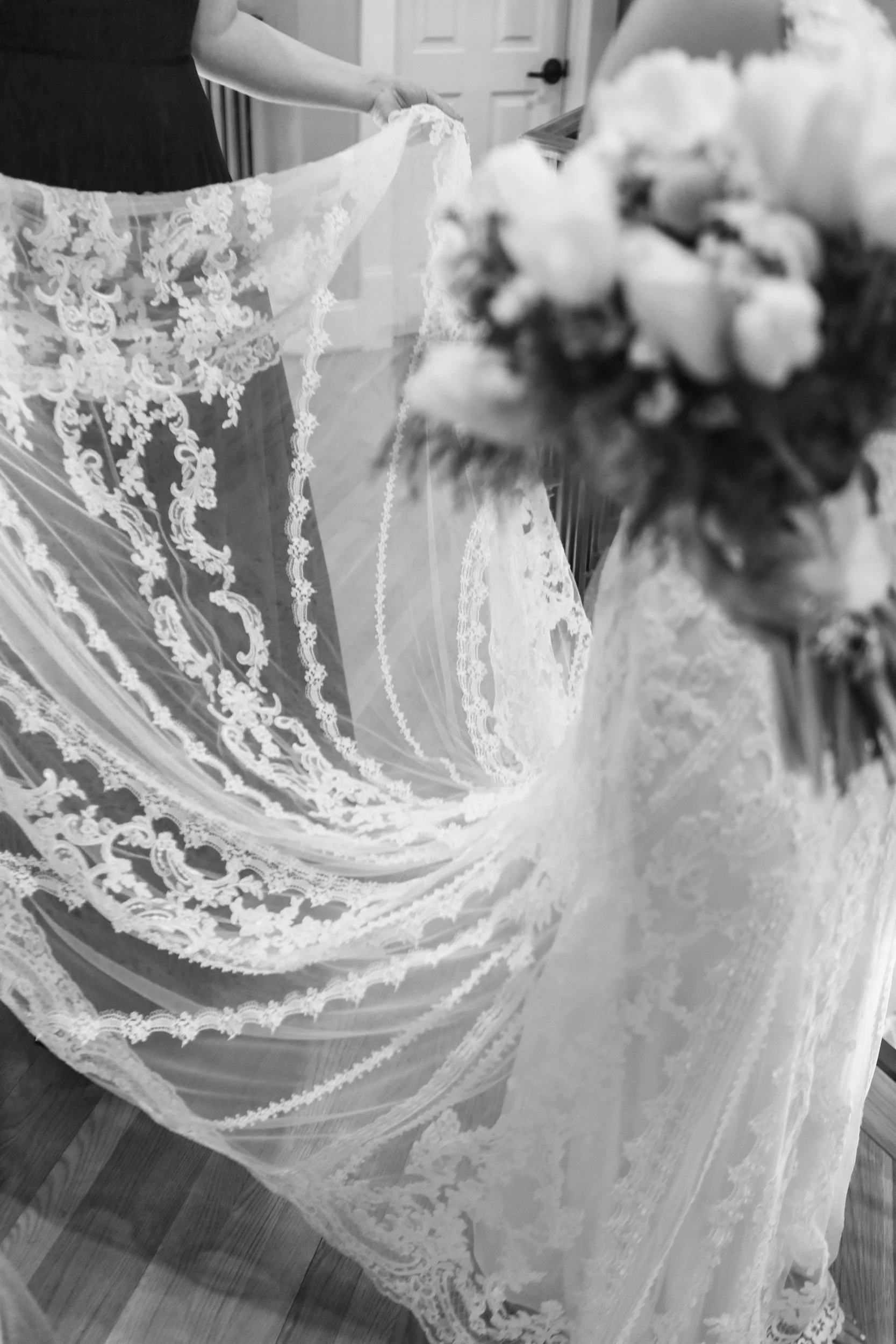 Black and white photo of a bride wearing a lace wedding dress, holding out her train, with a bouquet of flowers in the foreground.