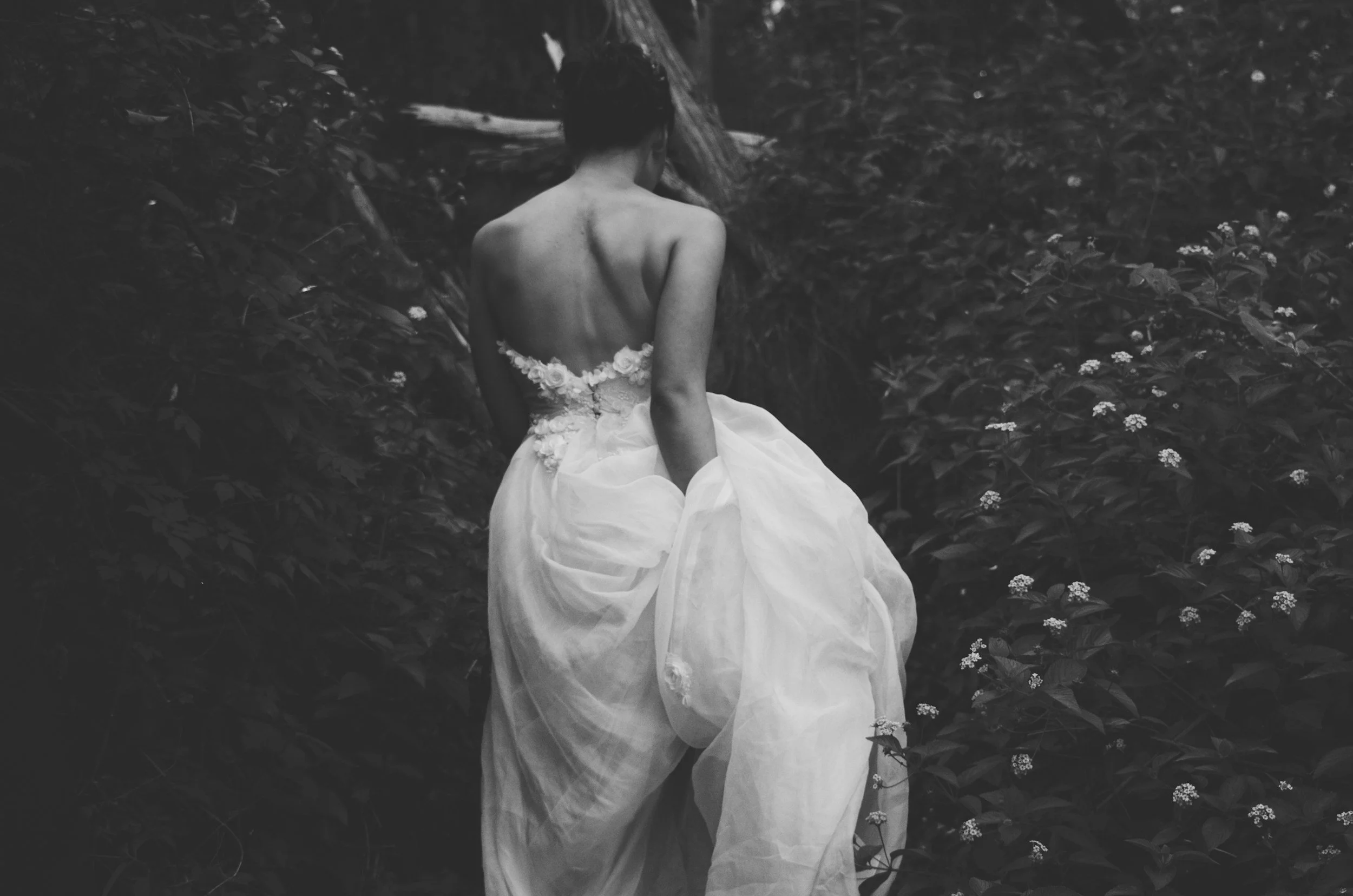 A woman in a strapless wedding dress walking through a garden of small flowers, viewed from behind.