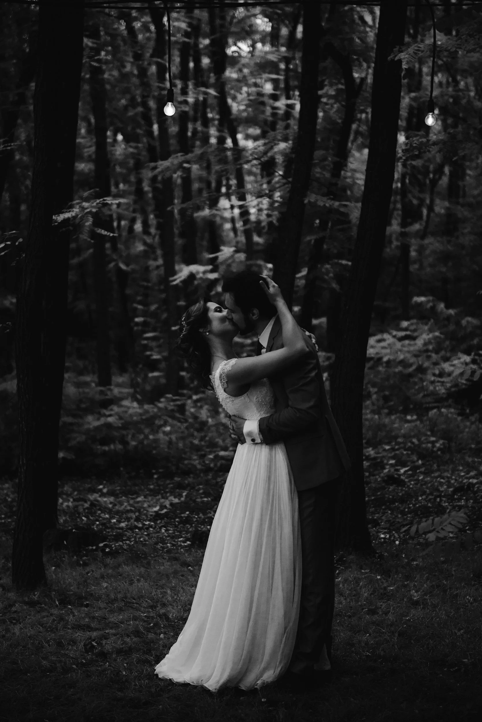 A black and white photo of a couple in wedding attire sharing a kiss in a forest, with string lights hanging overhead.