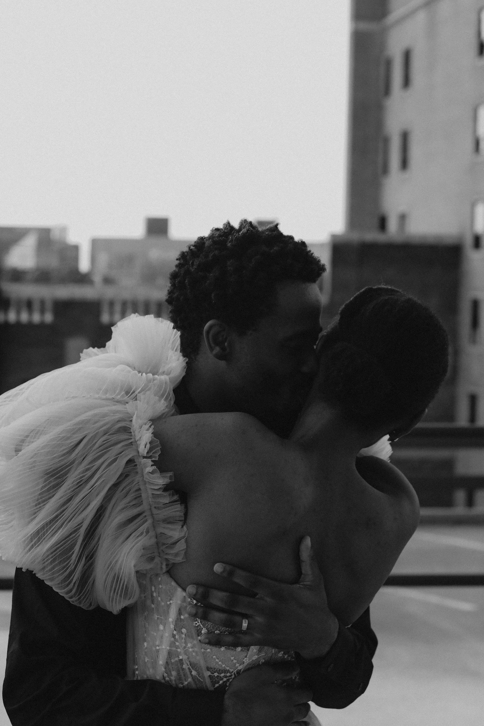 Two people kissing, one wearing a dress with ruffled fabric, in an urban setting in black and white.