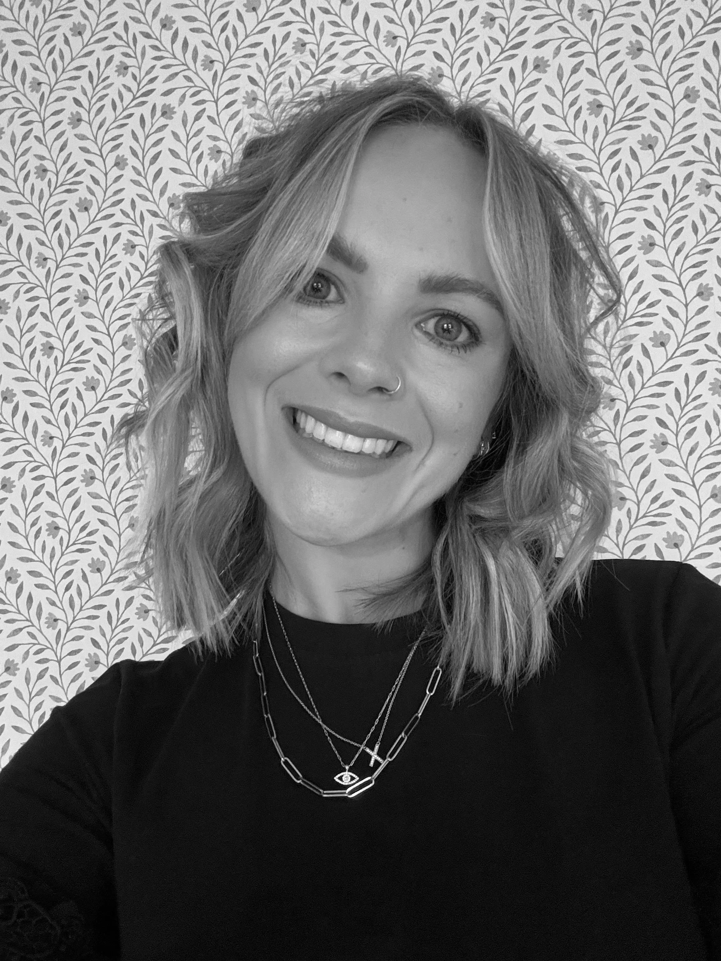 A smiling woman with wavy hair, wearing layered necklaces and a nose ring, in front of a patterned leafy wallpaper.