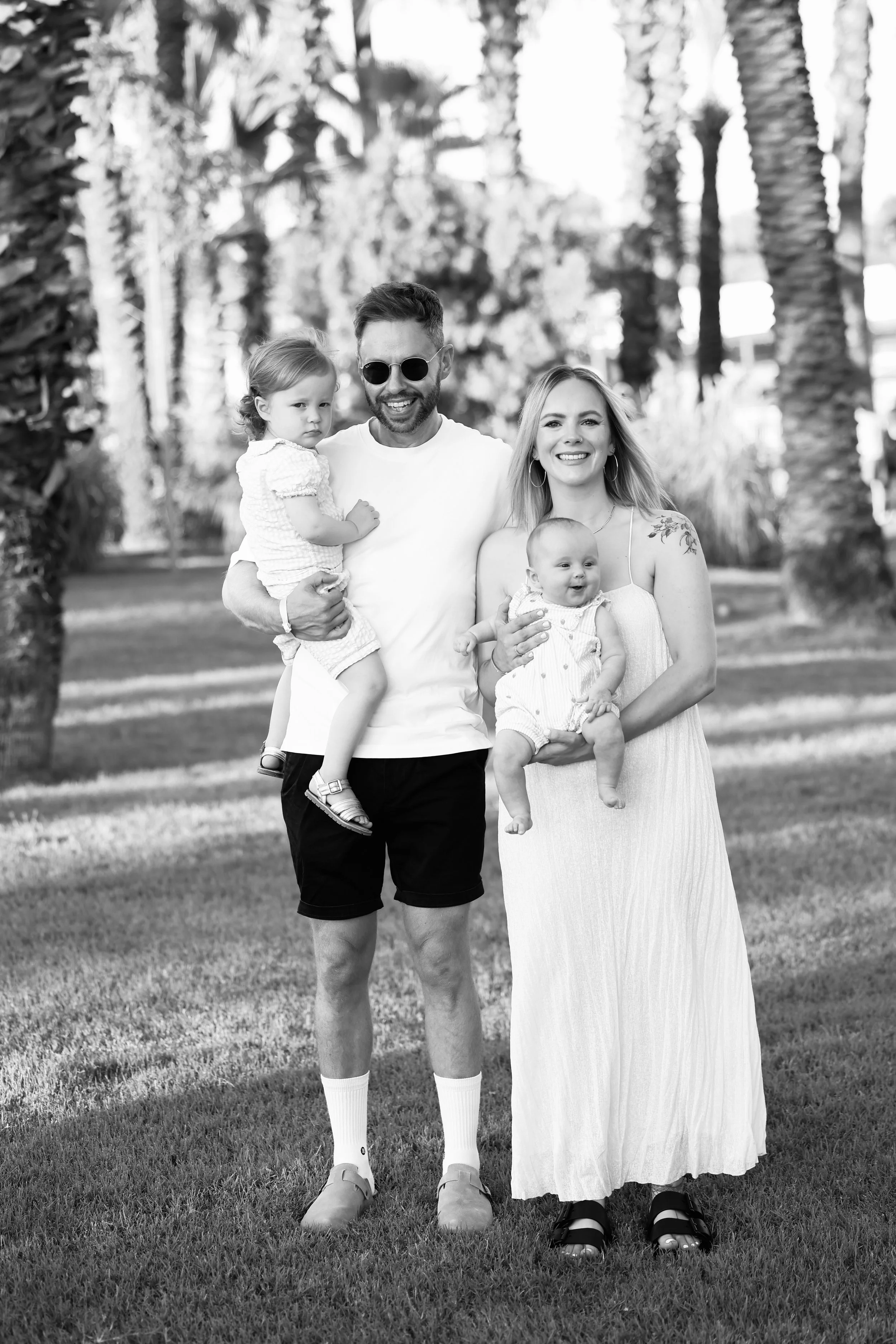 A smiling family of four outdoors in a park, with man holding a young girl and woman holding a baby, all dressed in light-colored summer clothing, surrounded by palm trees.