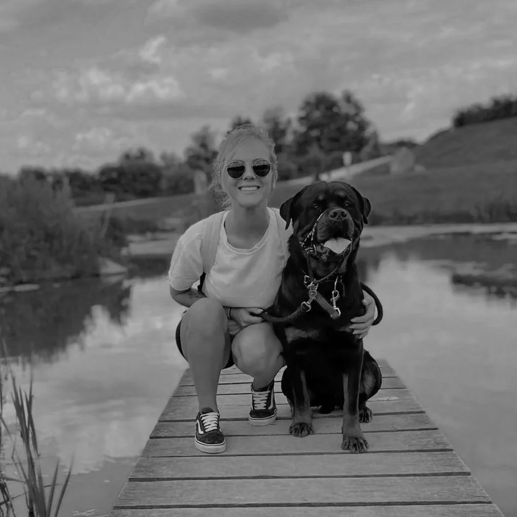 A woman wearing sunglasses, a white t-shirt, and sneakers kneels next to a large Rottweiler dog on a wooden dock beside a calm body of water. The woman is smiling, and the dog is sitting with its tongue out, wearing a leash and collar. The background shows trees and a partly cloudy sky.