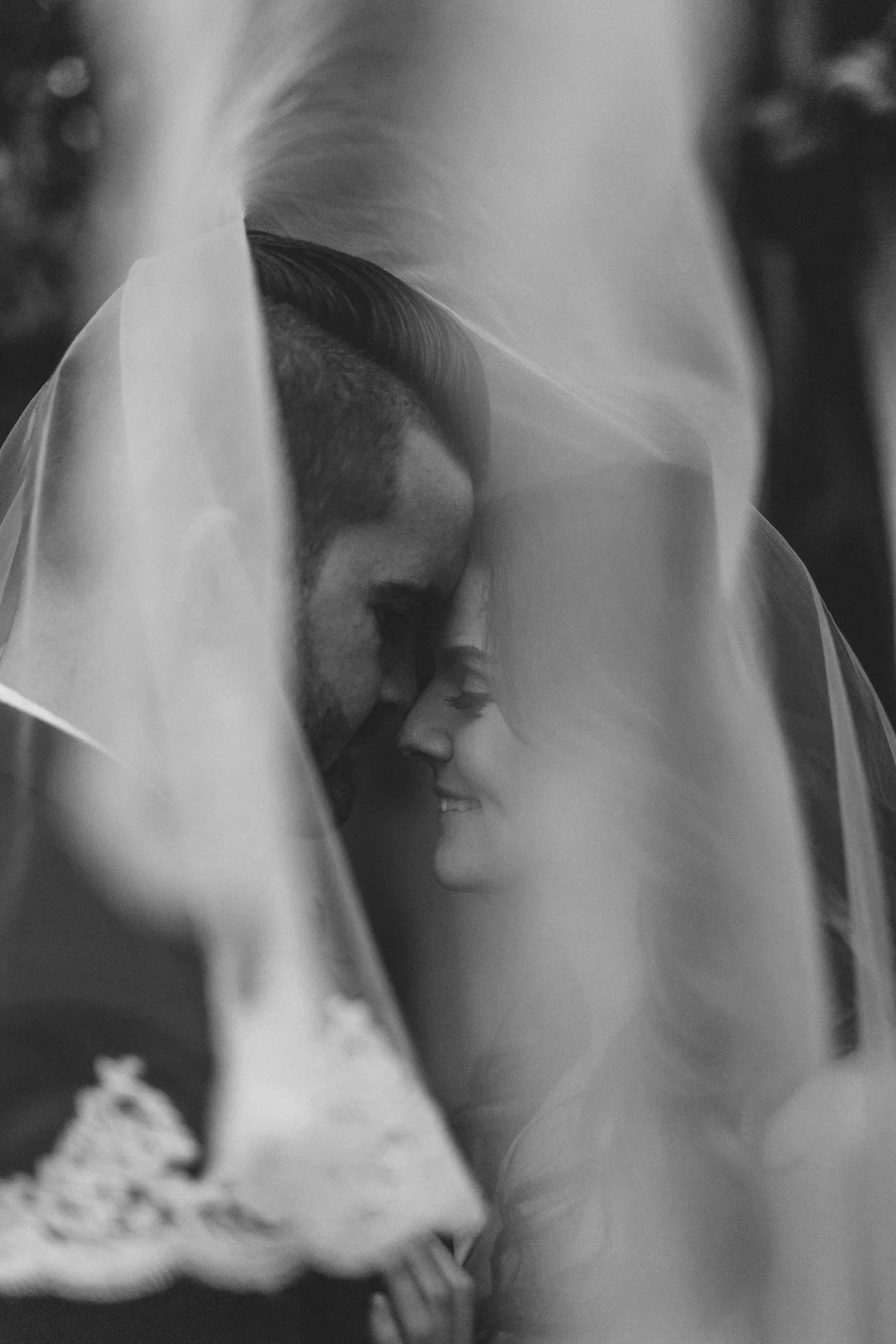 A close-up black and white photo of a man and woman with foreheads touching, both smiling, partially covered by a veil.