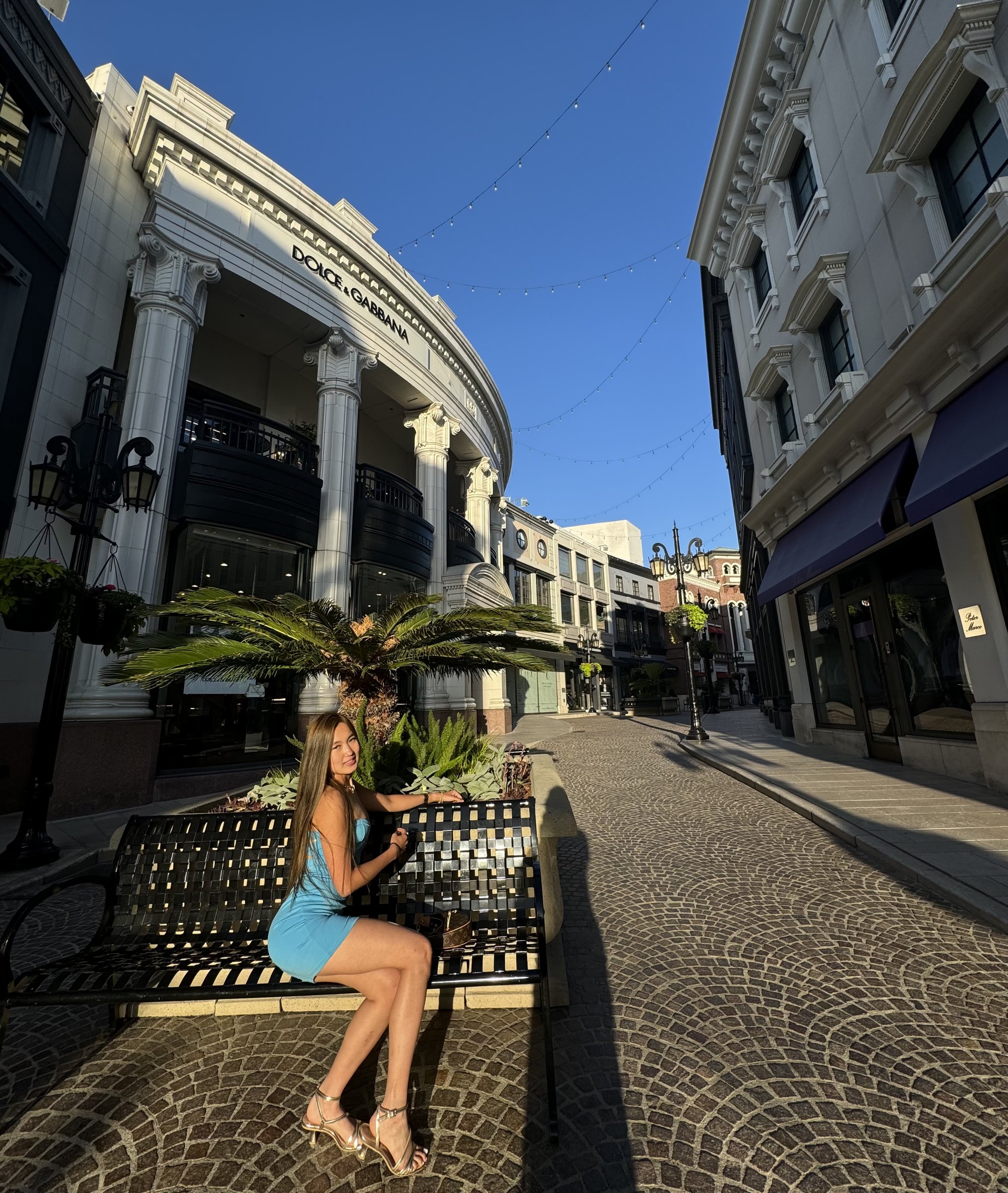 A woman in a blue dress and high heels sitting on a black metal bench next to a small palm tree and plants on a sidewalk in a shopping district with luxury stores, including Dolce & Gabbana, under a clear blue sky.