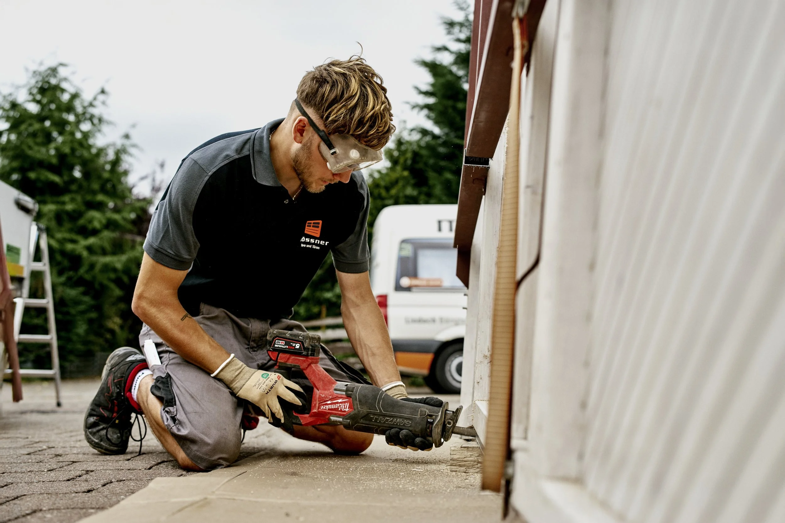 Ein junger Mann arbeitet auf einer Baustelle, nutzt eine Stichsäge, kniet auf dem Boden und trägt Schutzbrille, Handschuhe und Arbeitskleidung.
