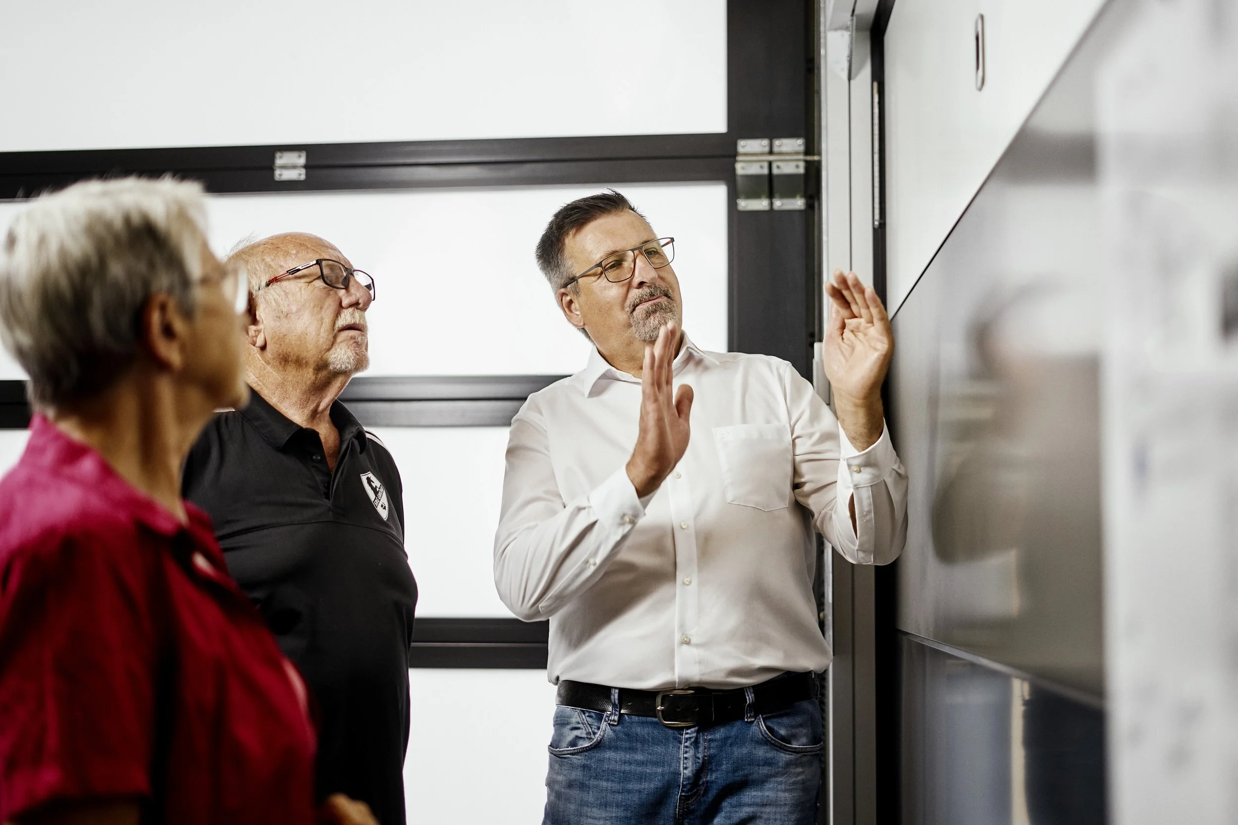 Drei Erwachsene im Gespräch vor einer Tafel, eine Person erklärt etwas mit Handgesten, im Hintergrund eine helle Wand.