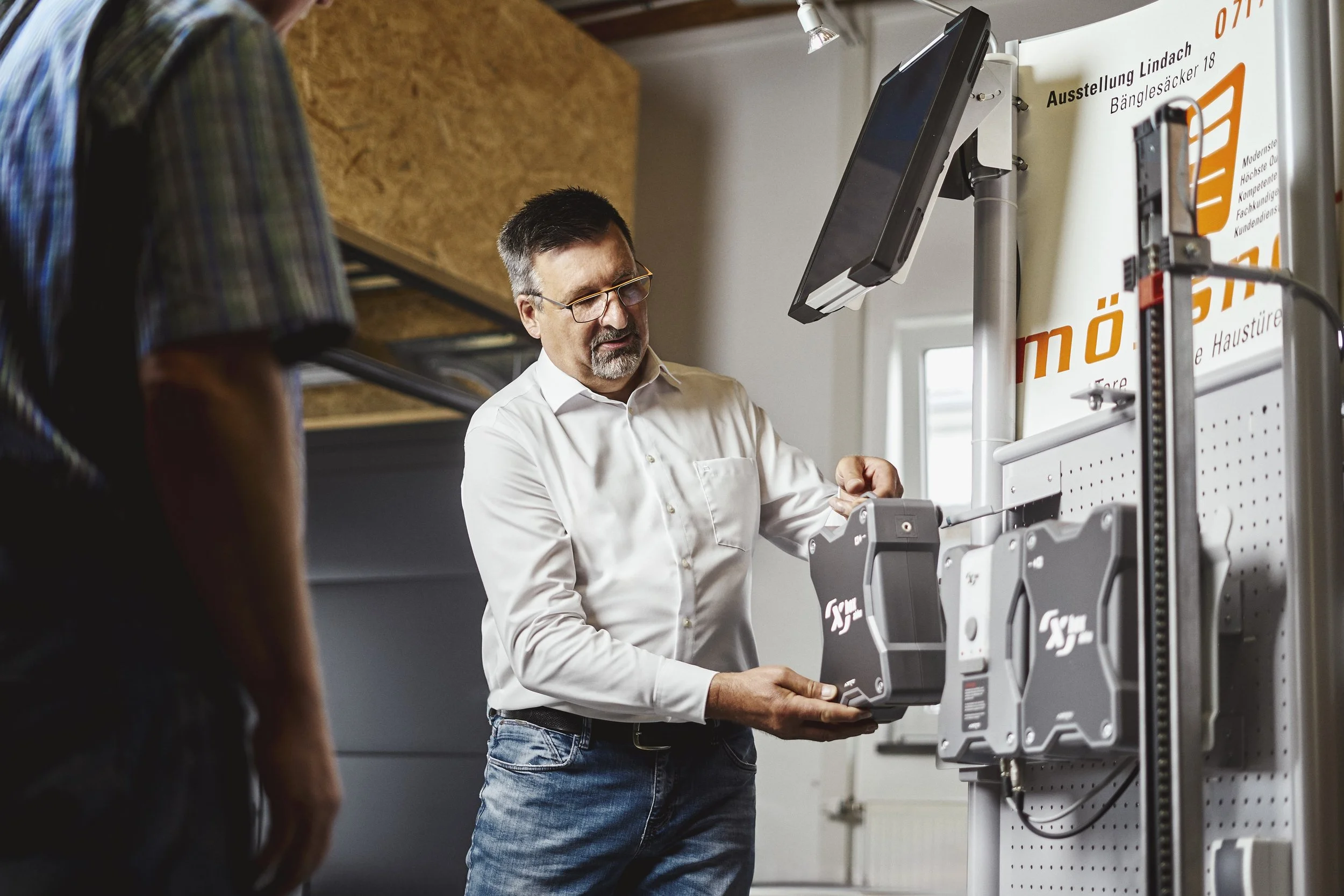 Ein Mann im weißen Hemd prüft eine elektronische Türsteuerung an einer großen metallischen Tür in einem Laden oder Ausstellungsraum.