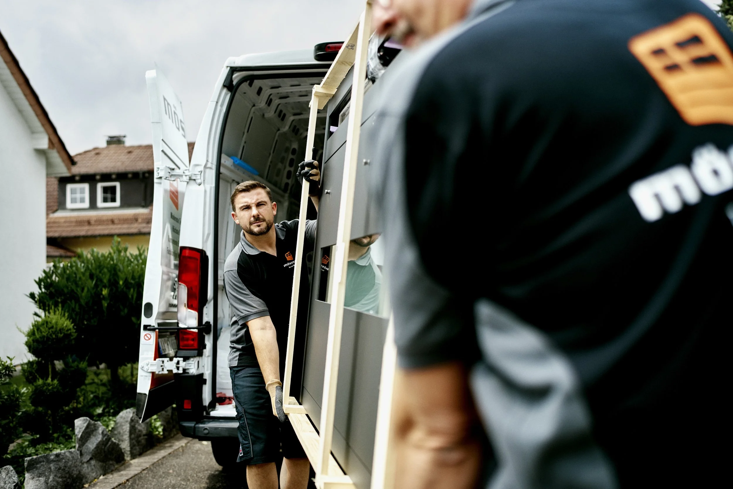 Zwei Männer arbeiten an einem Projekt im Freien, beim Transportieren eines Holzrahmens in einem weißen Lieferwagen, während ein Mann im Vordergrund teilweise sichtbar ist.