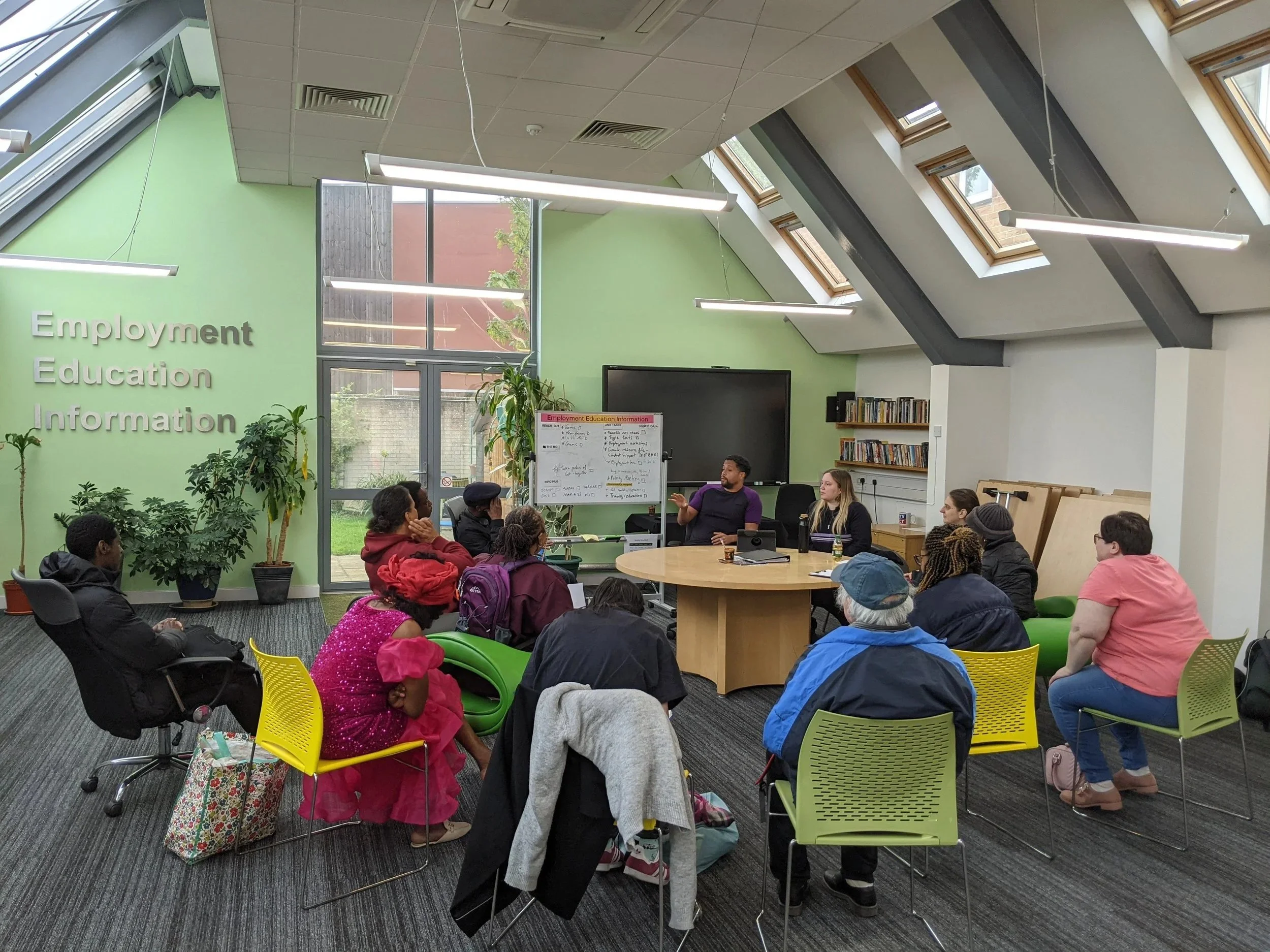 A group of diverse people sitting around a circular table in a classroom or community center, listening to a man who is speaking. The room has large windows, a bright green wall with the words "Employment Education Information," and a whiteboard with notes. There are plants, bookshelves, and skylights in the room.
