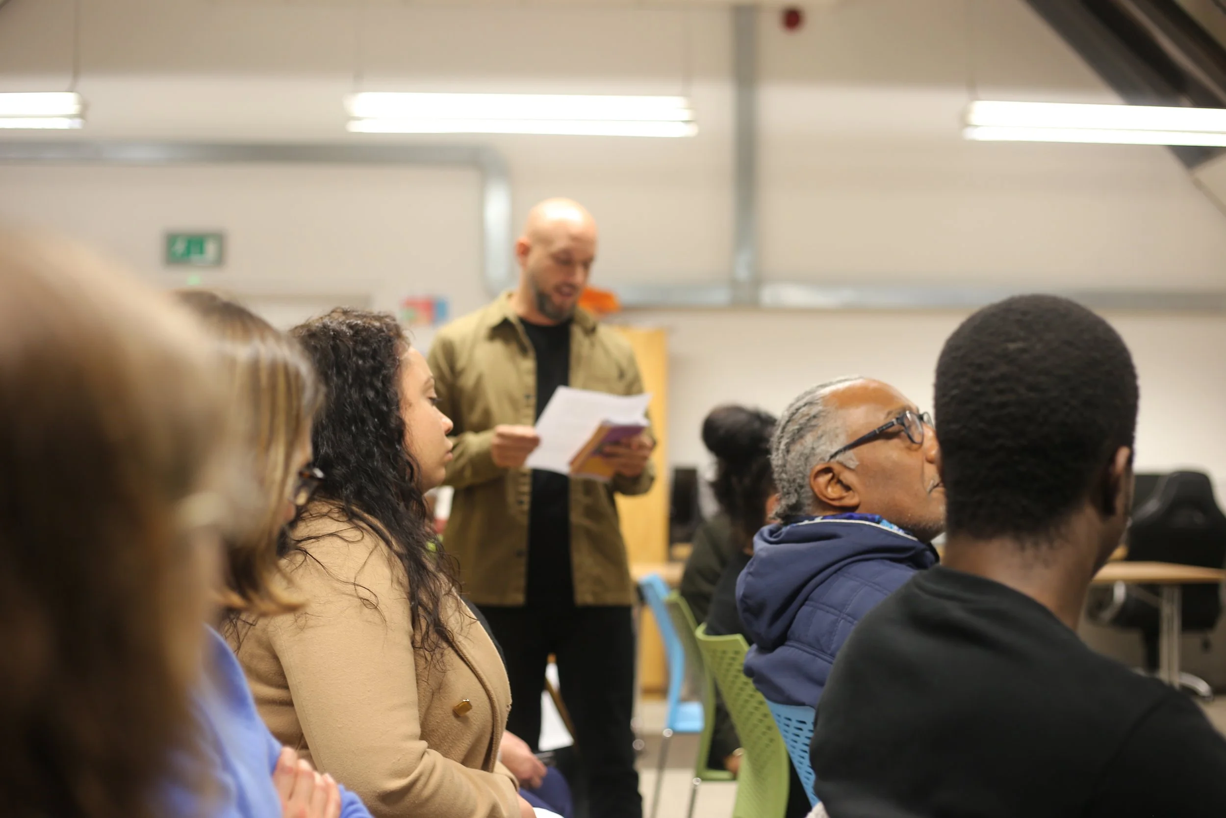 People sitting in a conference room or classroom, with a man standing and reading from a paper at the back.