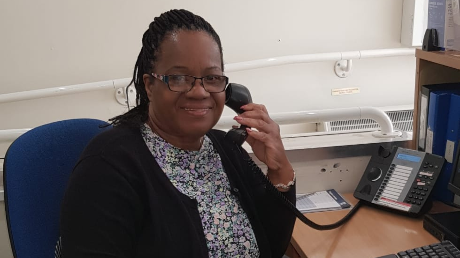 A woman with glasses talking on a landline phone at her desk in an office.
