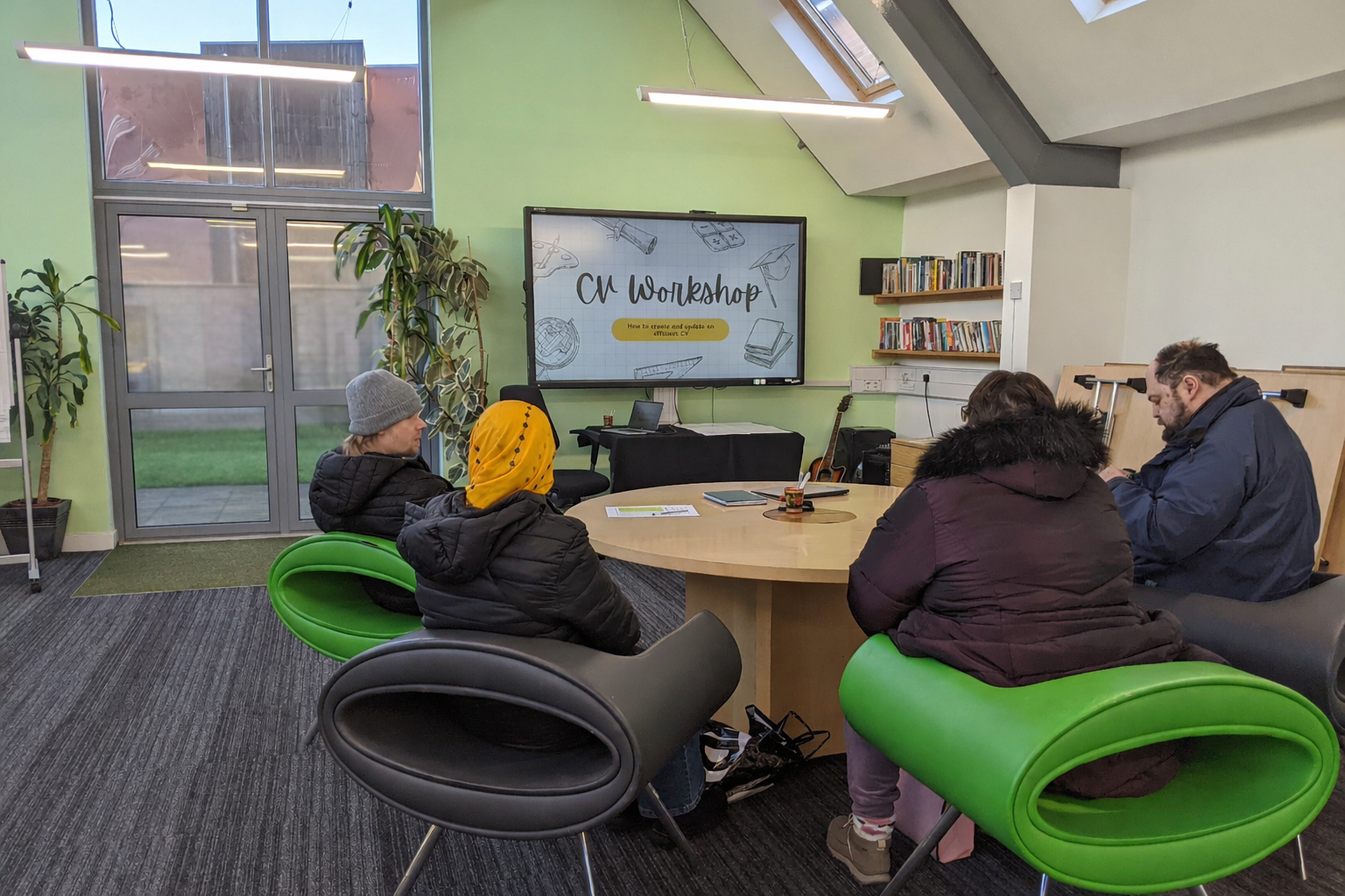 People attending a CV workshop in a modern room with green and white walls, large windows, and a digital screen displaying the workshop title.