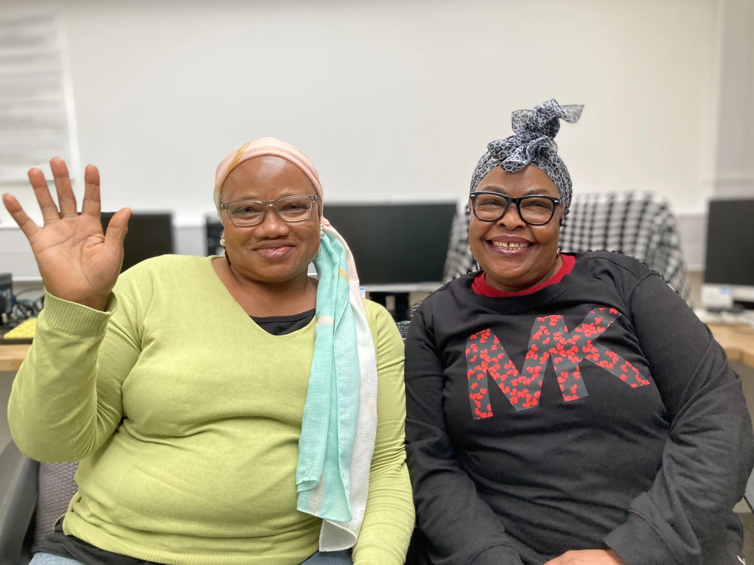 Two smiling women sitting together in an office, one waving, both wearing glasses and headscarves.