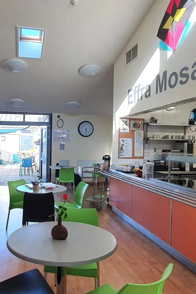 Interior of a cafe with green and black chairs, round tables, a vase with a single red flower, and a counter with food service equipment. Bright sunlight through an open door and window.