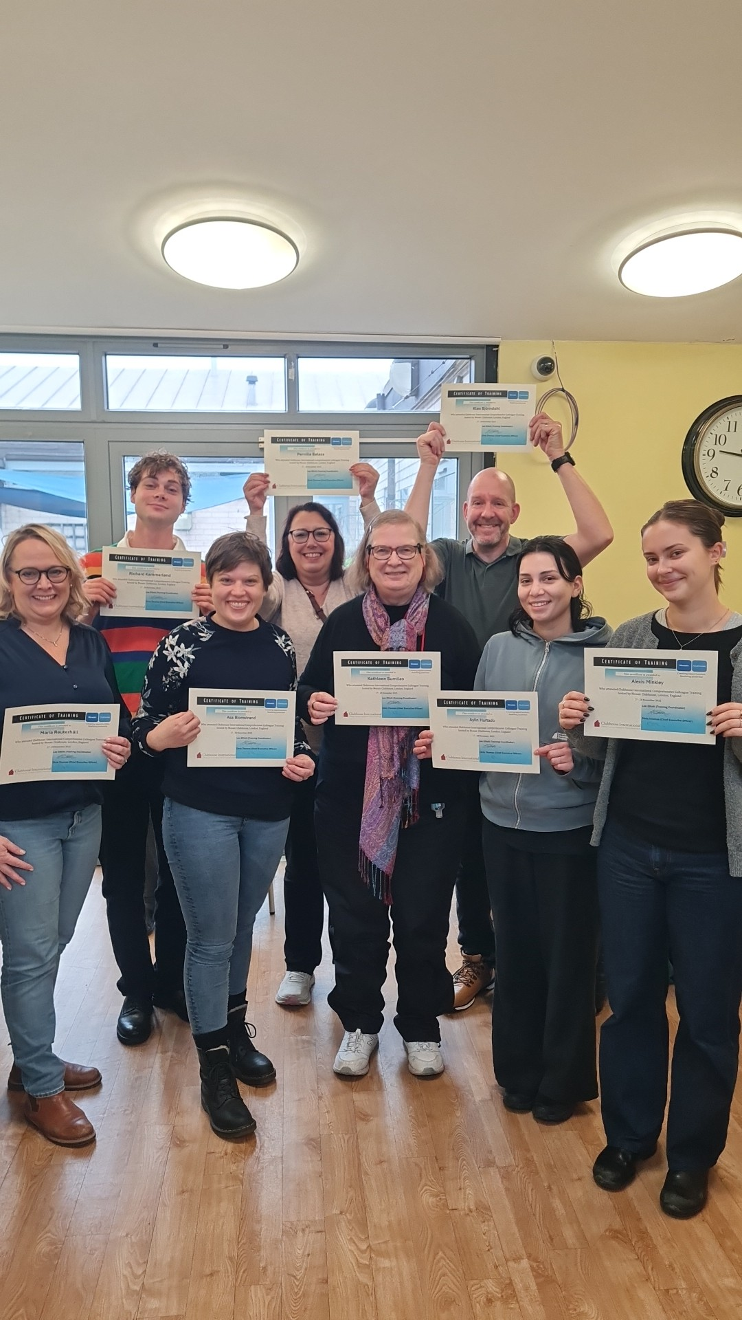 Group of nine smiling people holding certificates, standing in an indoor room with wooden floor, yellow walls, and windows at the back. Two people at the back are raising their certificates in the air.
