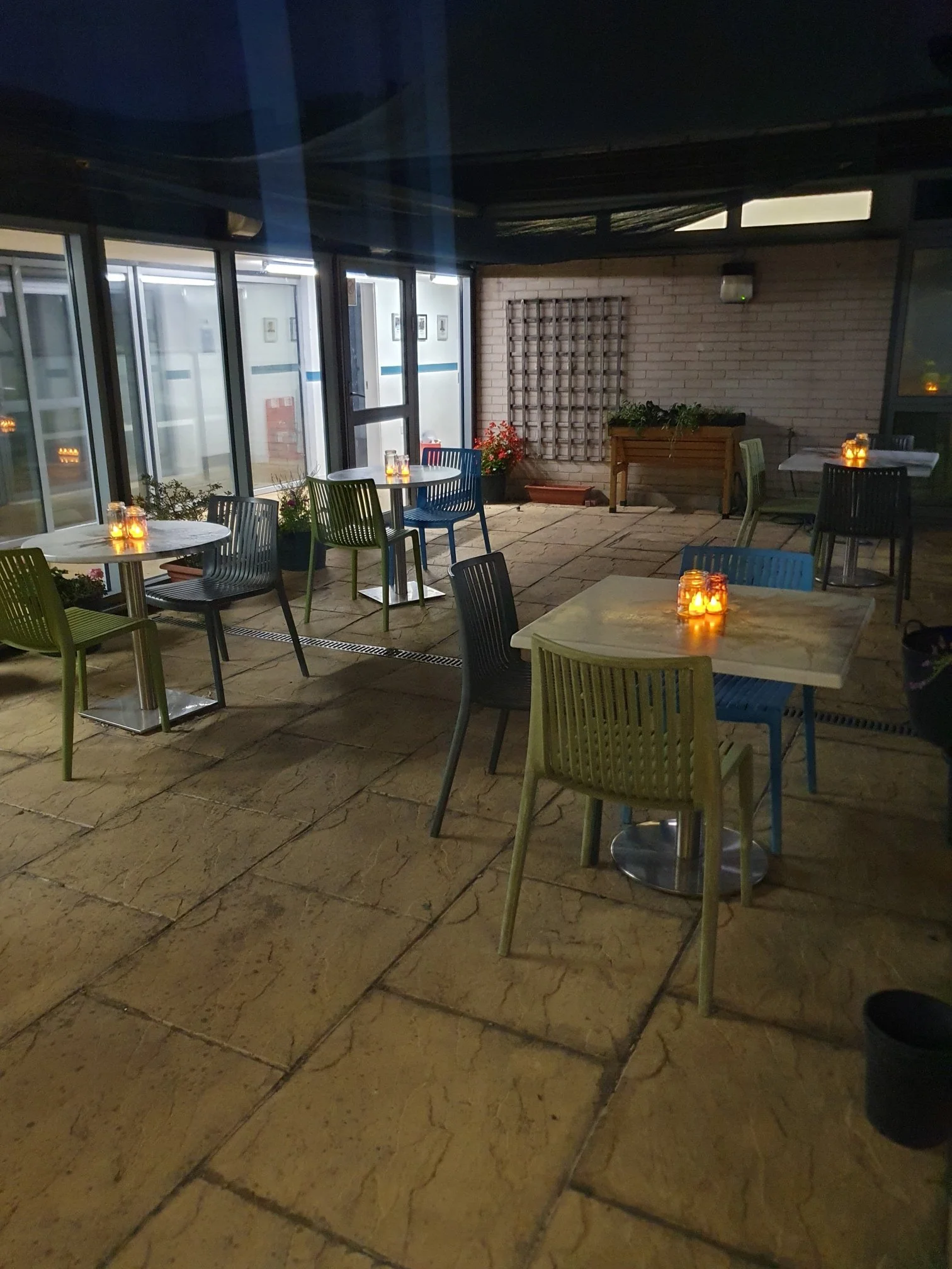Empty outdoor patio area with tables, chairs, lit candles, and potted plants at night.