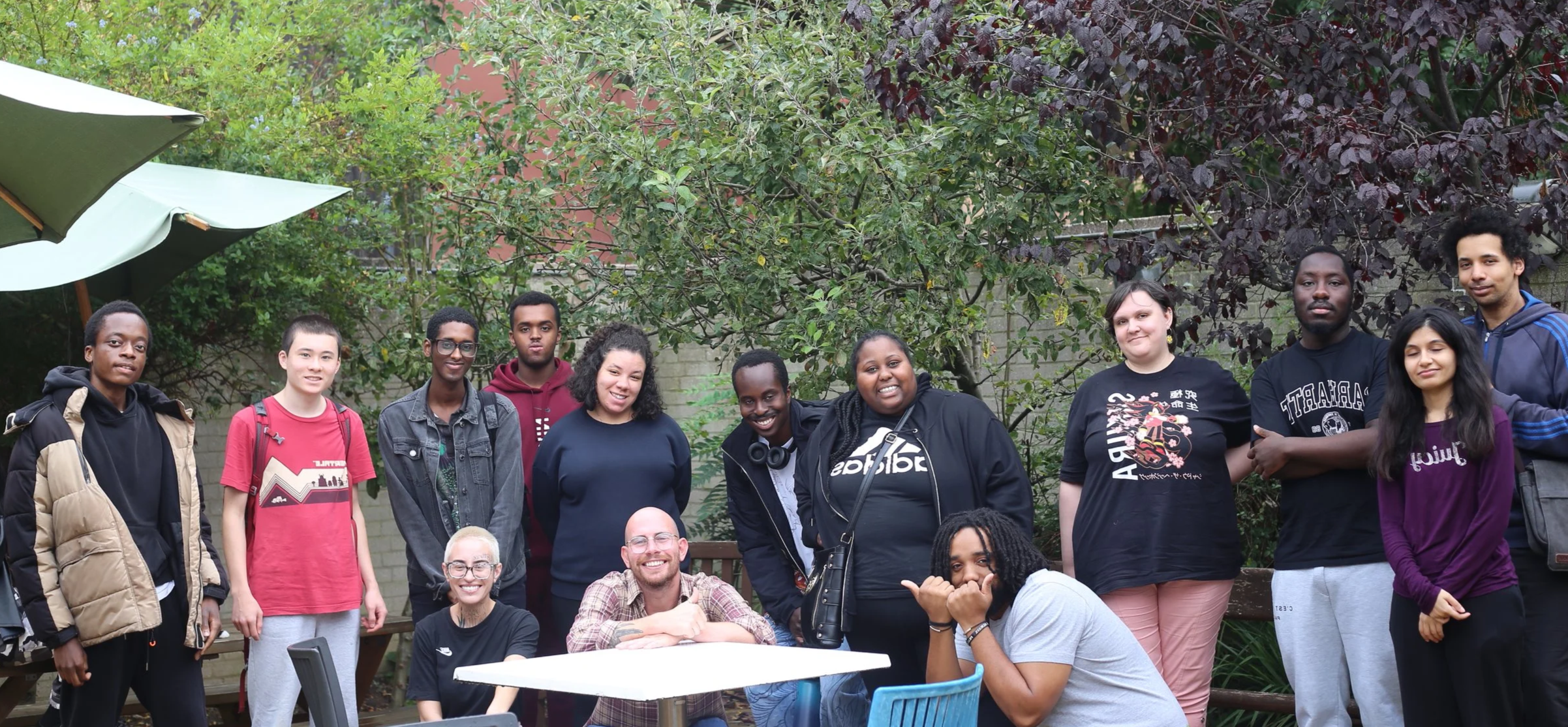 A group of diverse people posing outdoors in a garden area with trees and bushes, some standing and some sitting, smiling for the camera.