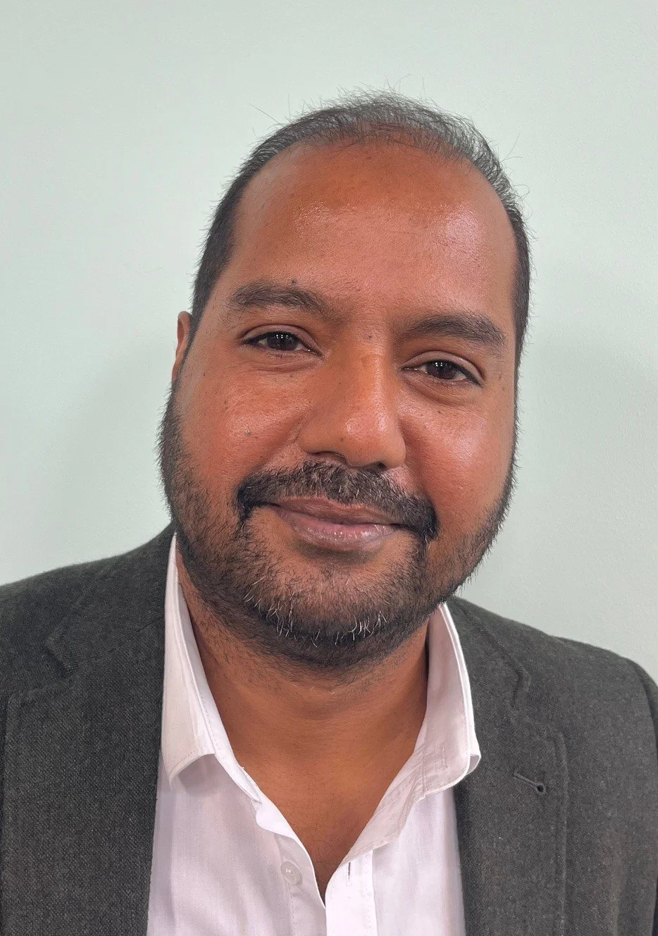 Close-up portrait of a man with short dark hair, a beard, wearing a dark blazer and white shirt, smiling softly against a plain light background.