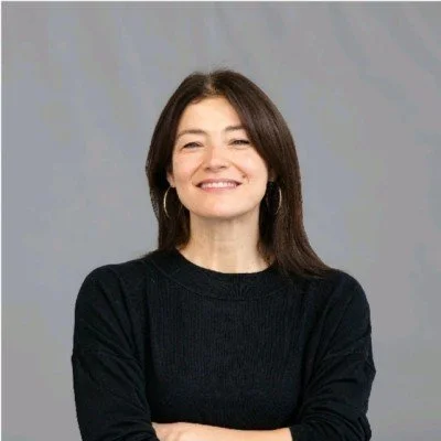 A woman with shoulder-length brown hair, wearing a red and white patterned sweater, sitting at a desk, smiling, writing on a yellow and white document with a green and white pen. There are papers and a computer on her desk in an office with light green walls.