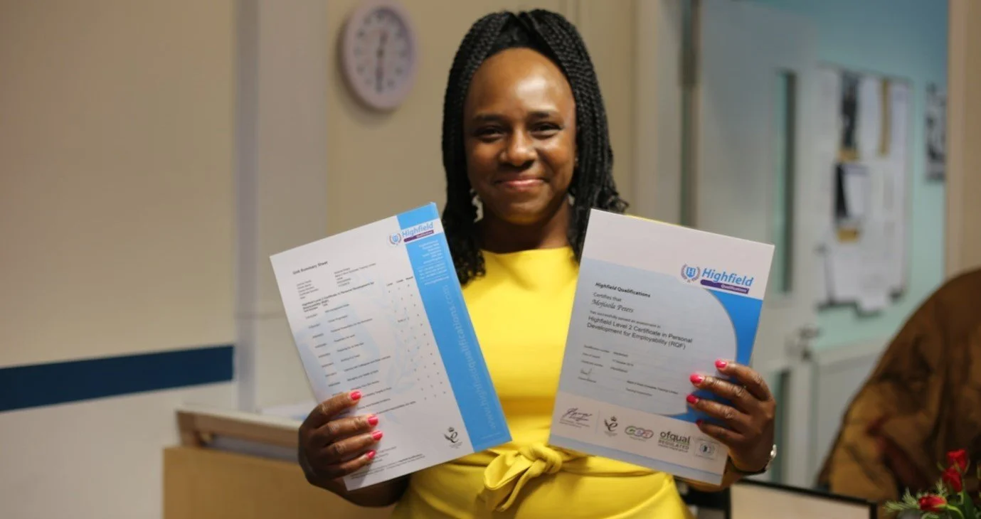 Woman smiling, holding two certificates from Highfield Qualifications in an indoor setting, with a wall clock in the background.