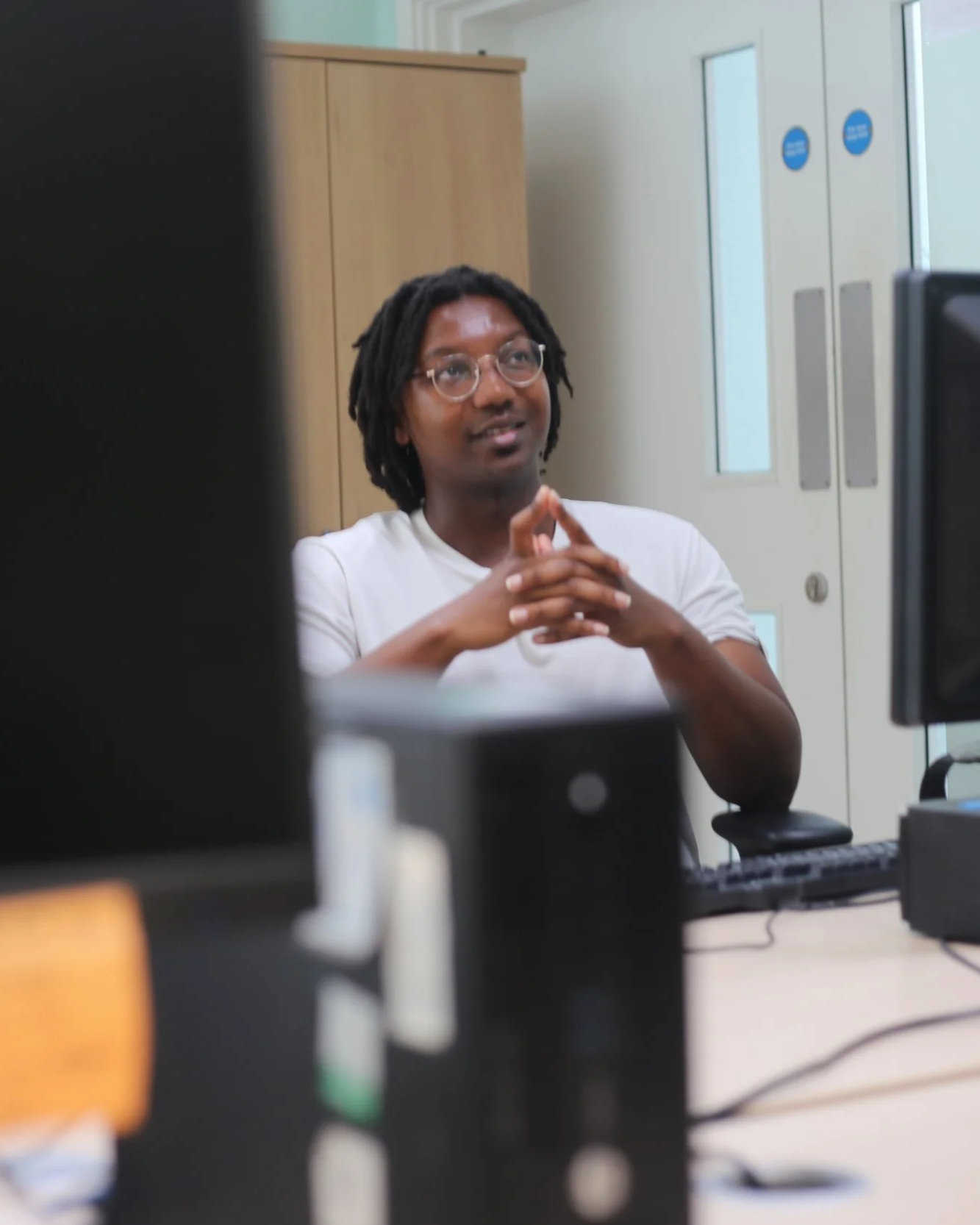 A man with glasses and dreadlocks sitting at a desk in front of a computer, with a serious expression, clasping his hands together. Privacy lockers and a window are in the background.