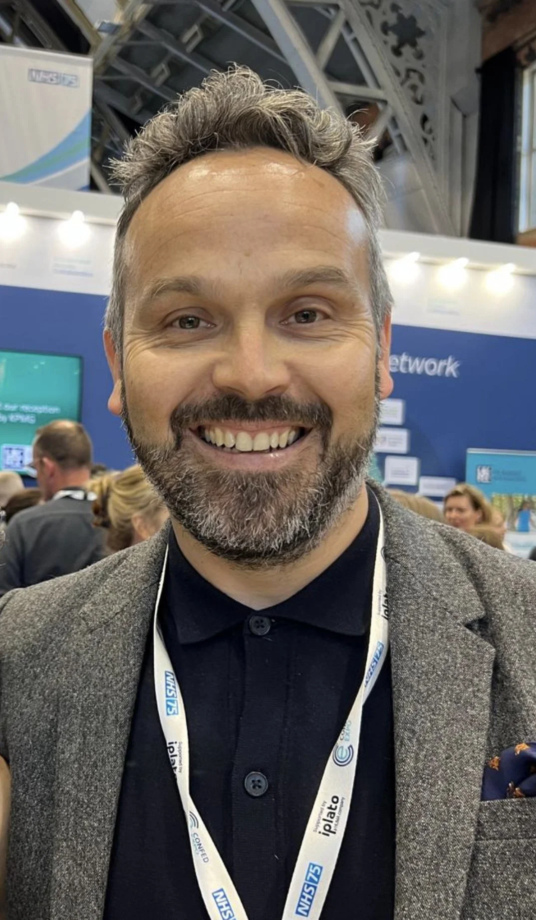 A man with a beard and gray hair smiling at a conference or event, wearing a black shirt and a gray blazer, with a lanyard around his neck, in a crowded exhibition hall.