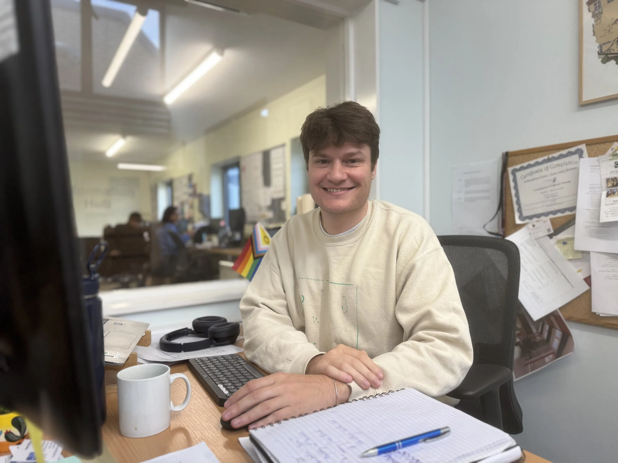 A man smiling, sitting at a desk with a cup, notebook, and headphones. Office environment with colleagues working in the background.