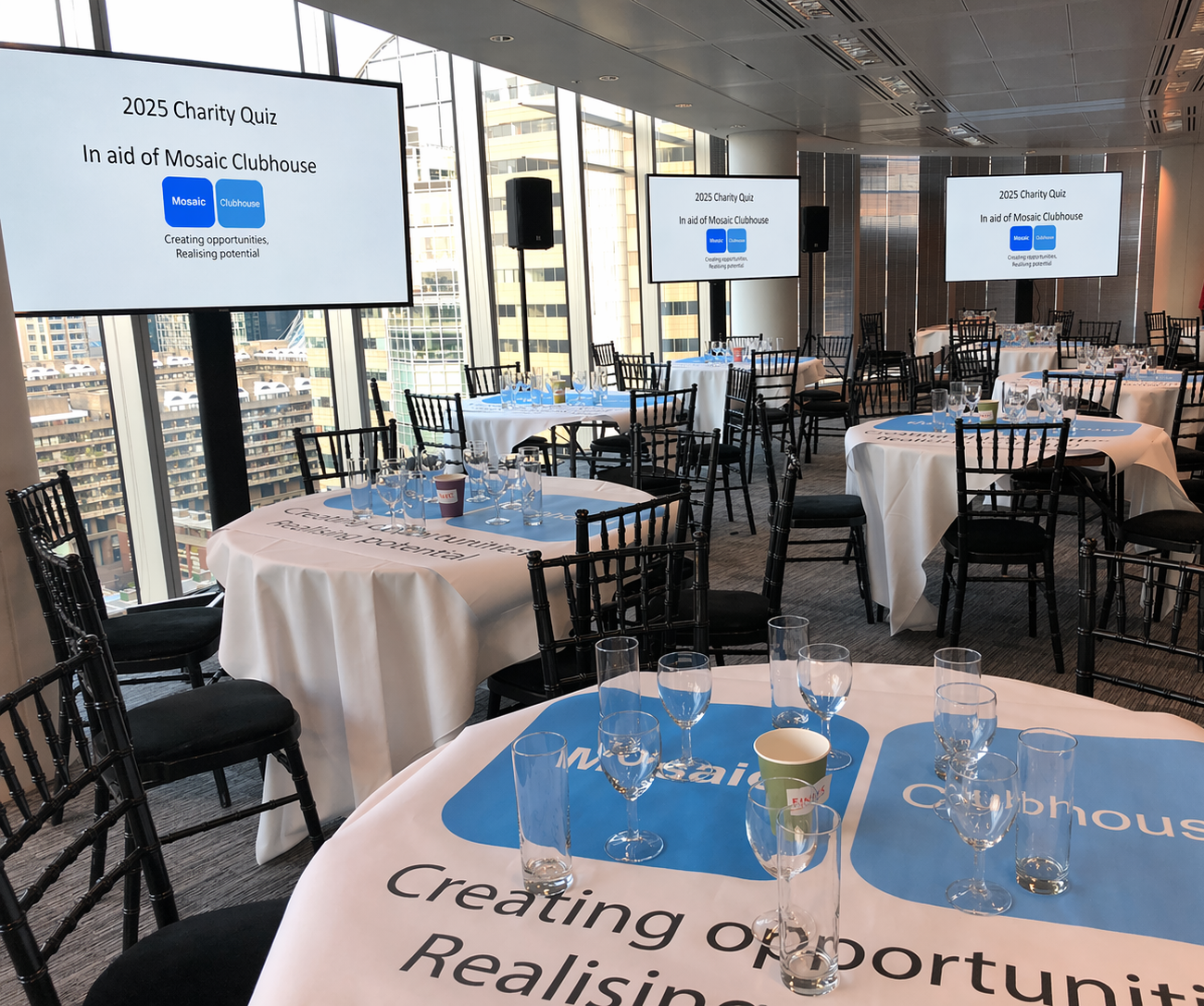 Conference room set up for the 2025 Charity Quiz in aid of Mosaic Clubhouse, with round tables covered in white tablecloths, each with multiple glasses, a coffee cup, and blue and white branding on the table covers. Large screens display event information, and the room has large windows showing a cityscape of high-rise buildings.