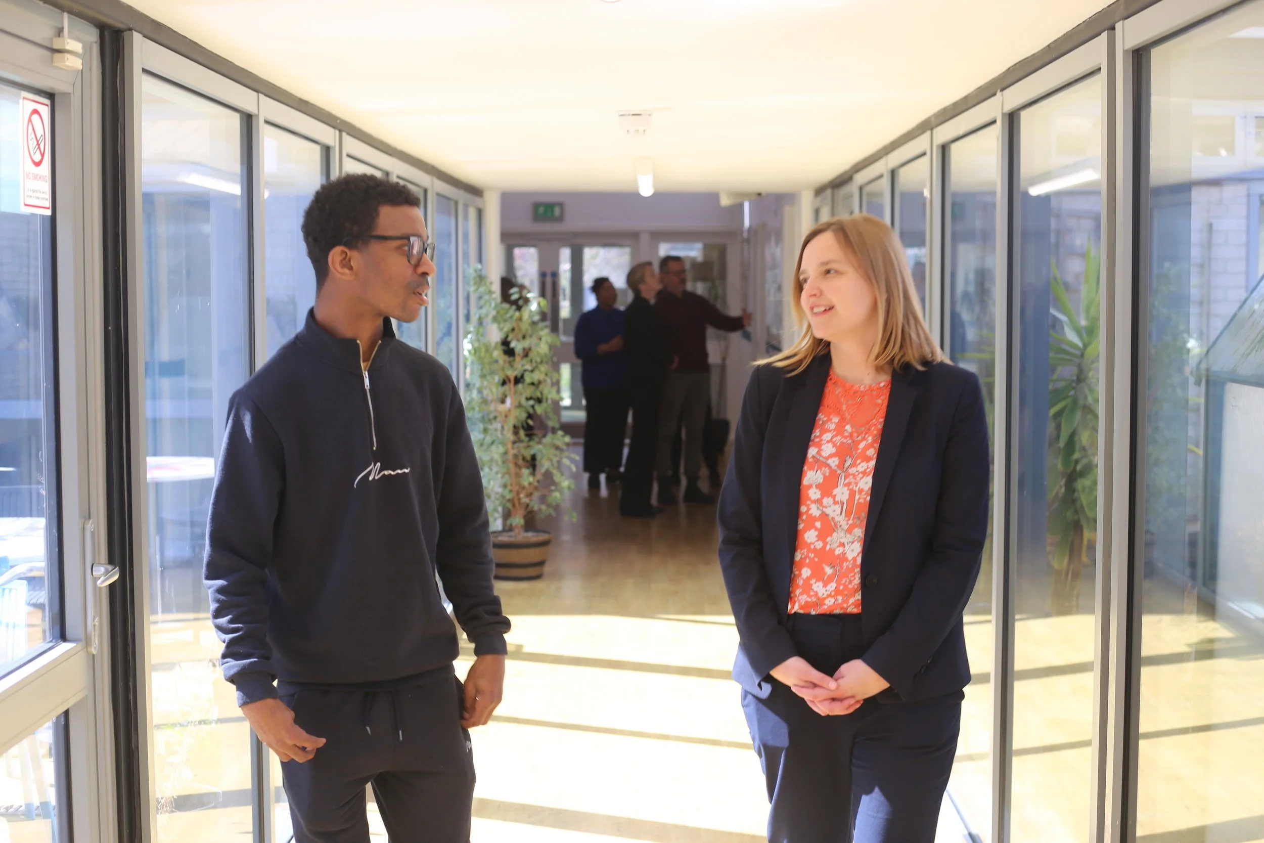 A man and a woman are talking and smiling in a bright hallway with large glass windows. In the background, other people are visible, and there are potted plants.