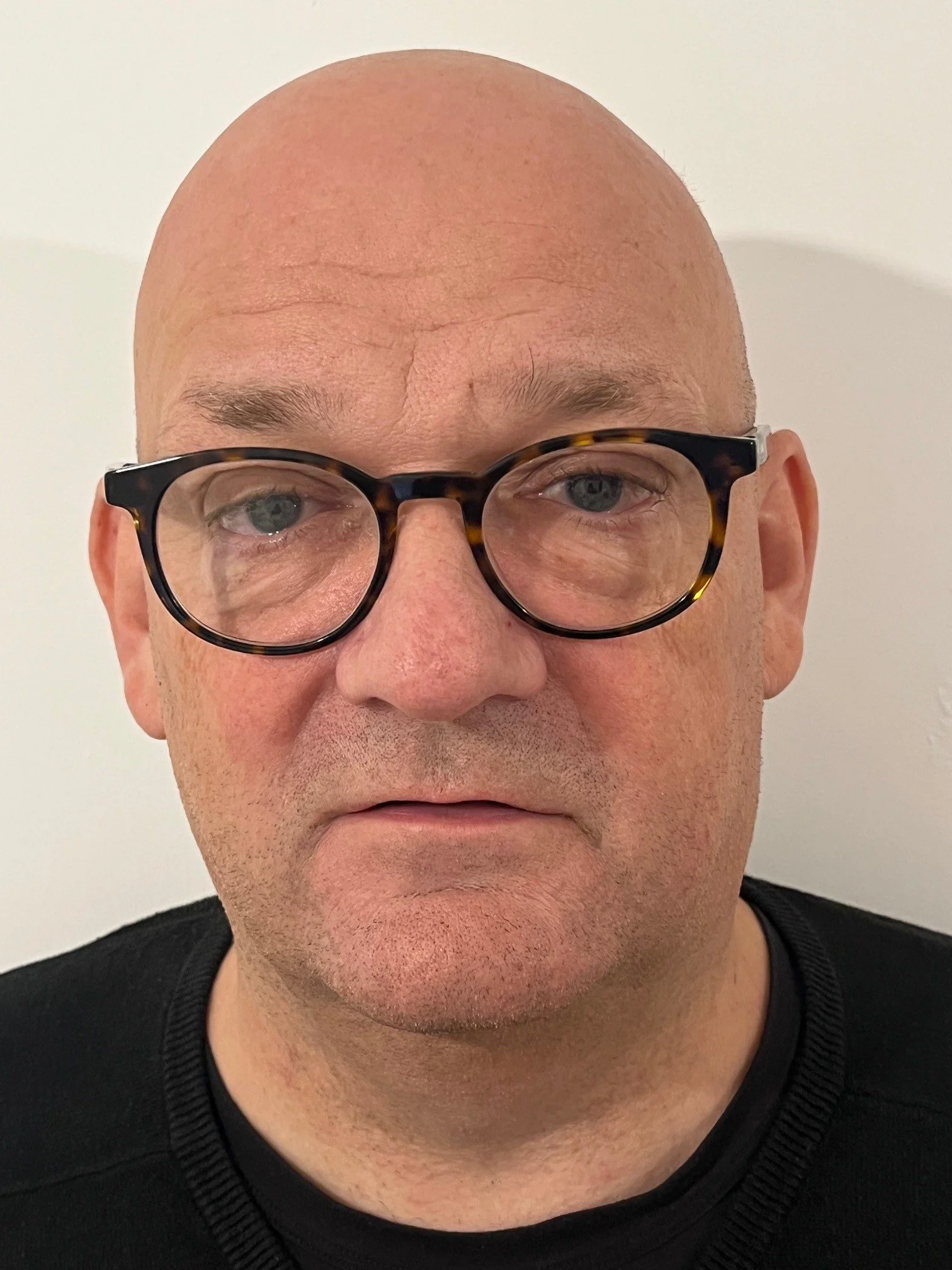 Close-up of a bald man wearing tortoiseshell glasses and a black shirt, looking directly at the camera against a plain white background.