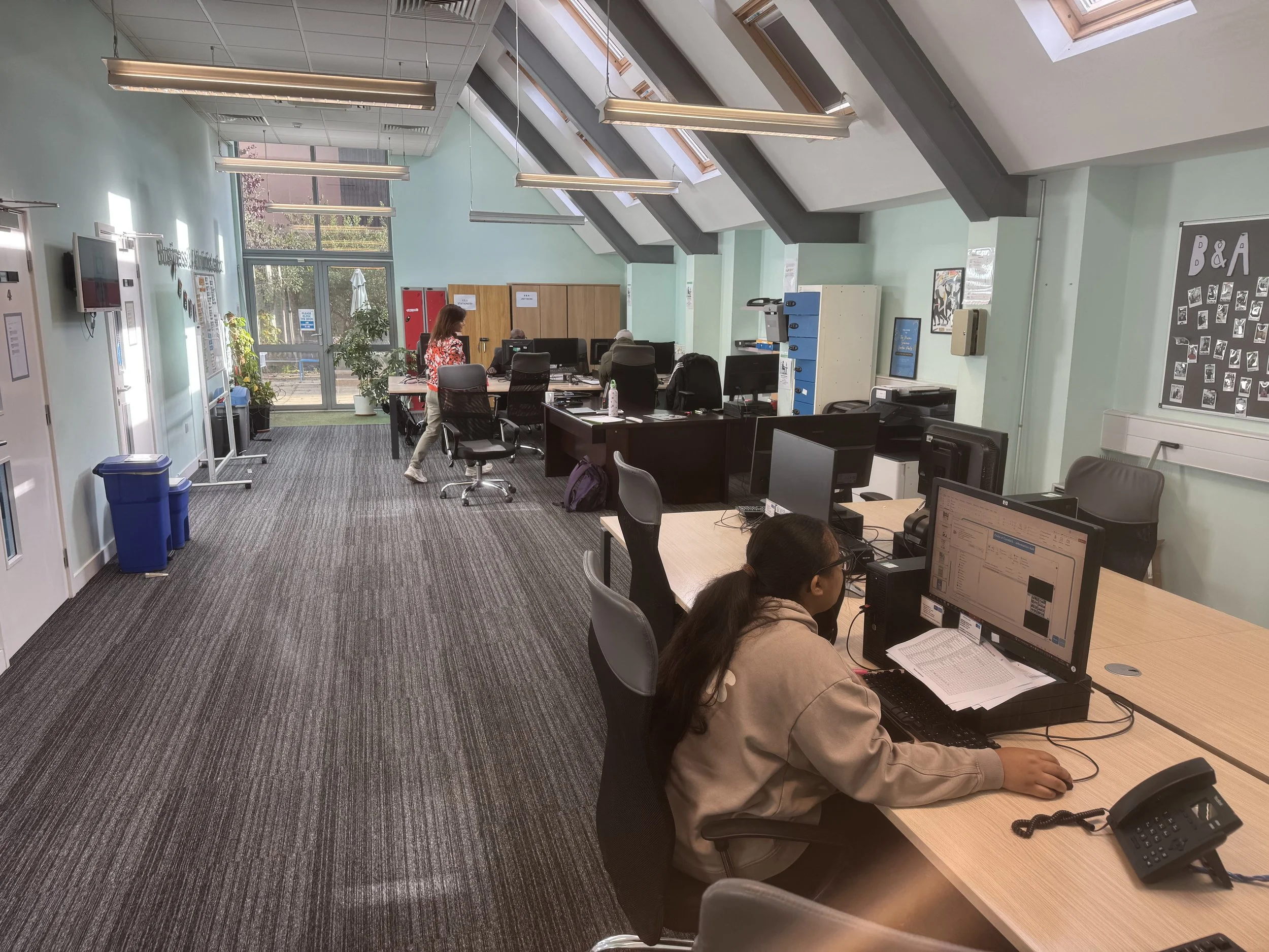 An office with several desks, people working at computers, and large skylight windows. There are plants near the entrance, a bulletin board on the wall, and various office supplies.