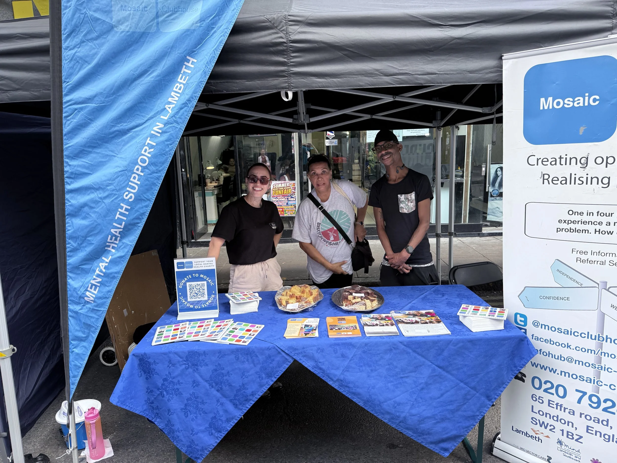 A booth at an outdoor event with three smiling people standing behind a table covered with a blue tablecloth. The table has brochures, flyers, and samples, with a banner displaying the website and social media information for 'Mosaic.' A large sign with text about mental health and a QR code is also on the table. The booth is under a black canopy tent with a blue flag on the side.