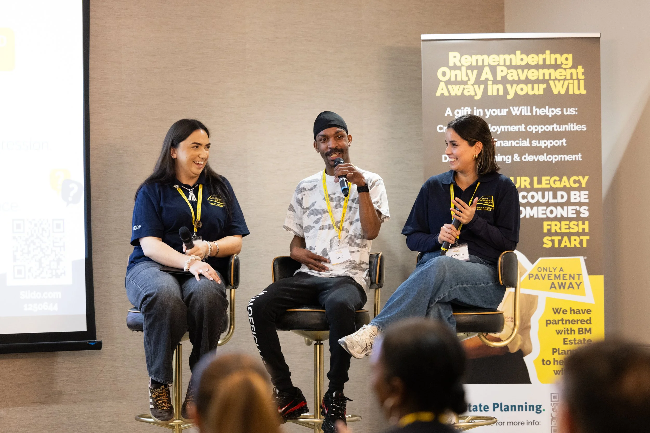 Three people are sitting on chairs on stage, speaking into microphones during a panel discussion. Two women and one man, all smiling, are engaged in conversation. Behind them is a banner with text about estate planning and a partially visible screen on the left.