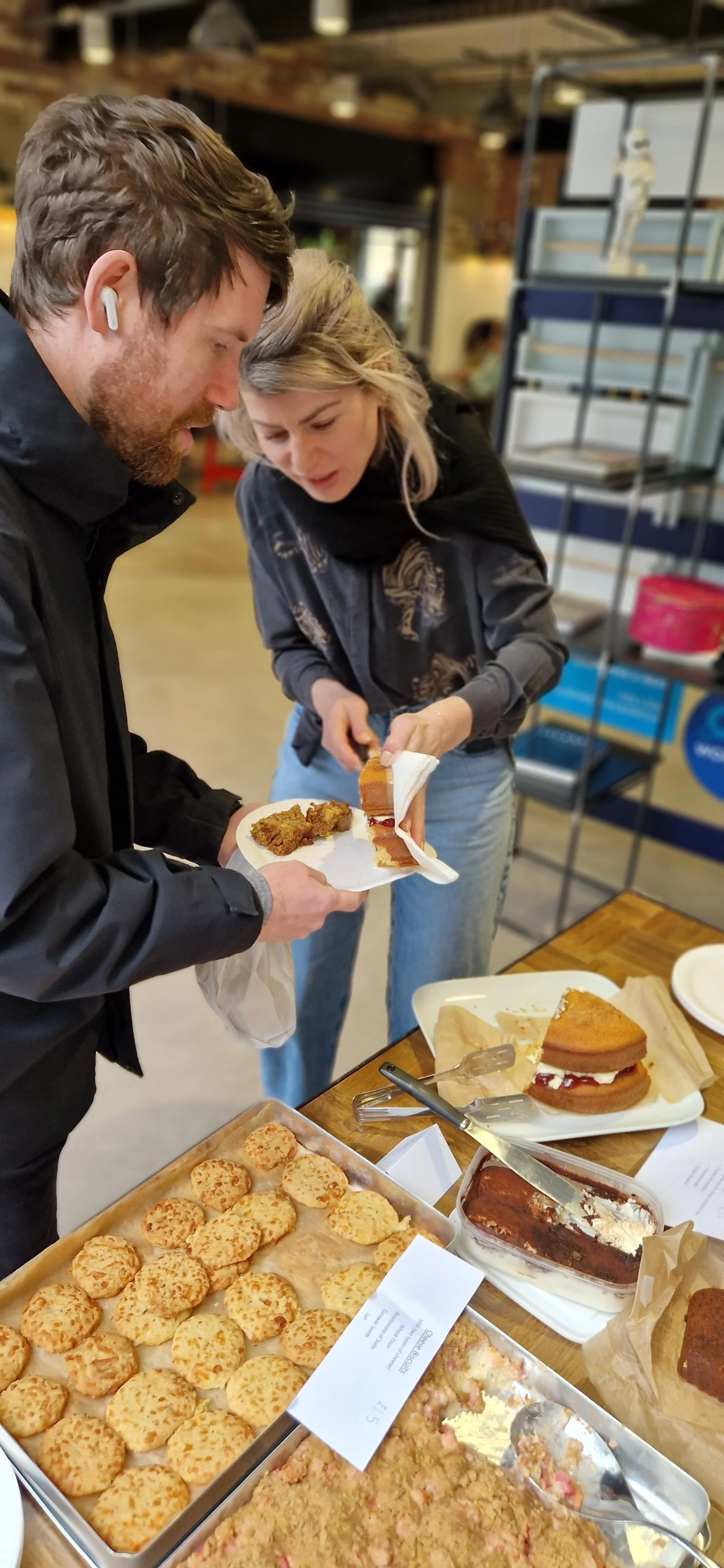 Two people at a table with various desserts, including cookies, cake slices, and a casserole dish, in a bakery or cafe setting.